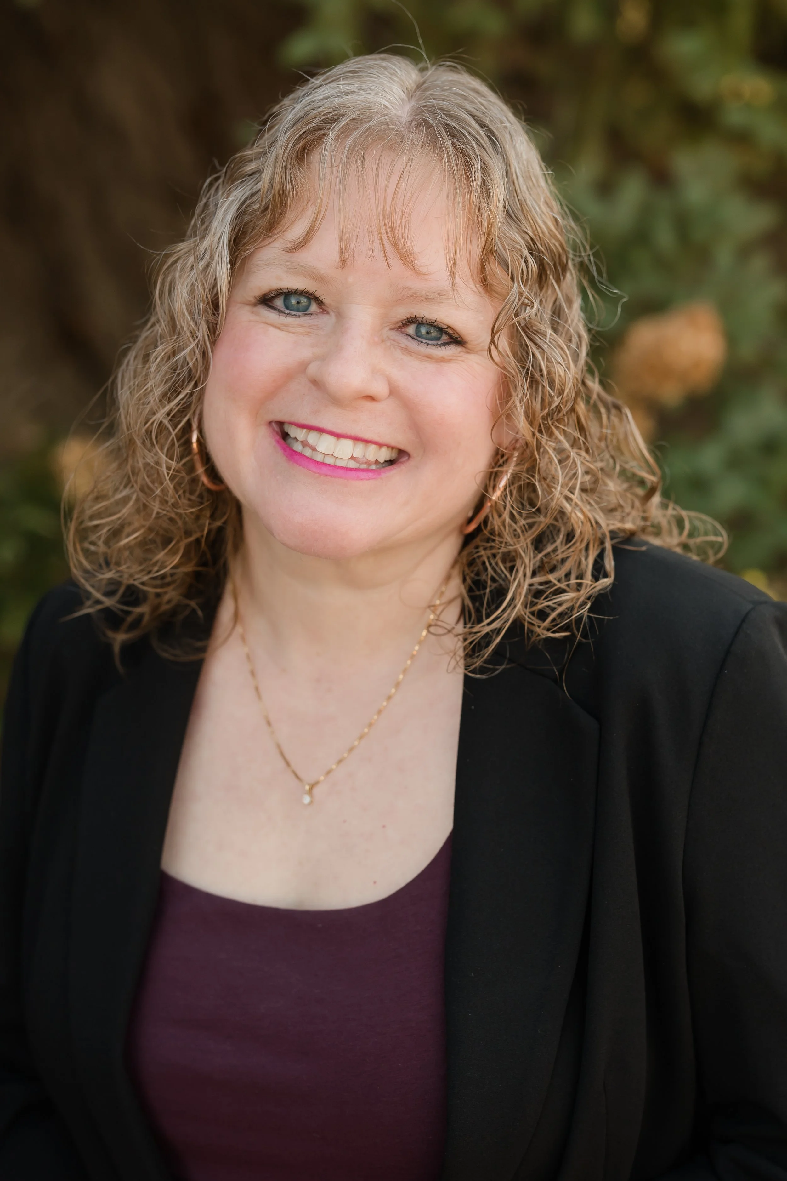 A woman with curly blonde hair wearing a black top, sitting on a beige sofa with a plant in the background.
