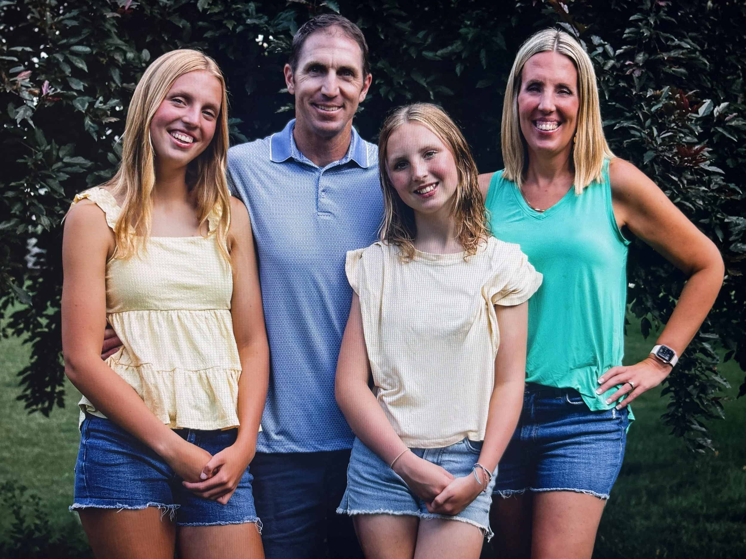 A family of five standing outdoors in front of a leafy tree, smiling at the camera. The family includes two women, one man, and two young girls, all dressed casually in summer clothing.