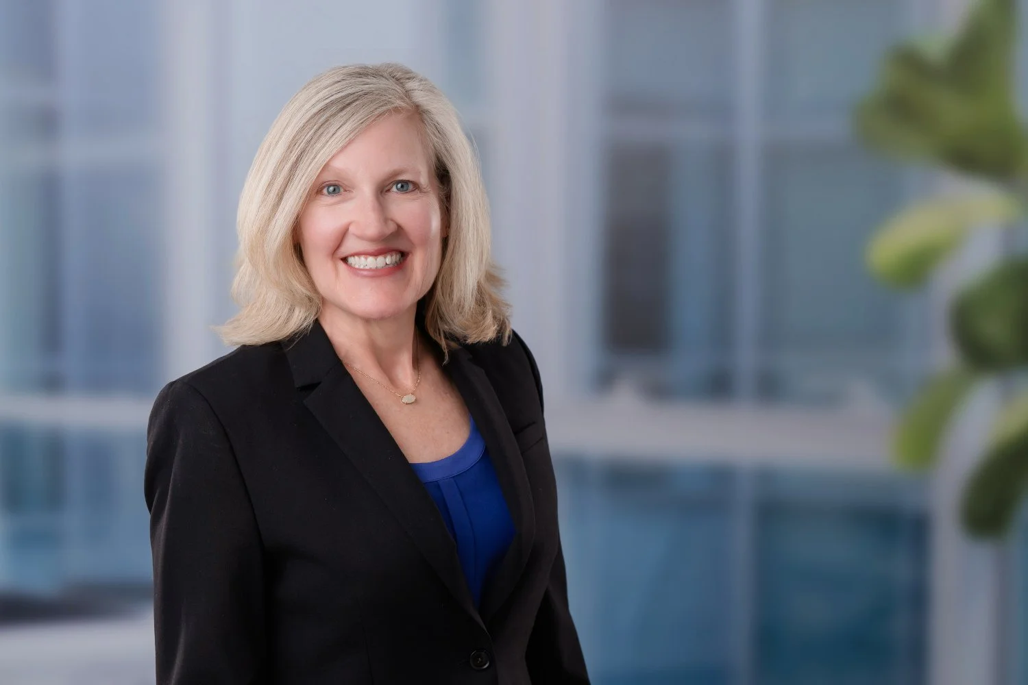 Headshot of a smiling woman with blonde hair, wearing a black blazer and a blue top, standing in a modern office with large windows and a blurred background.