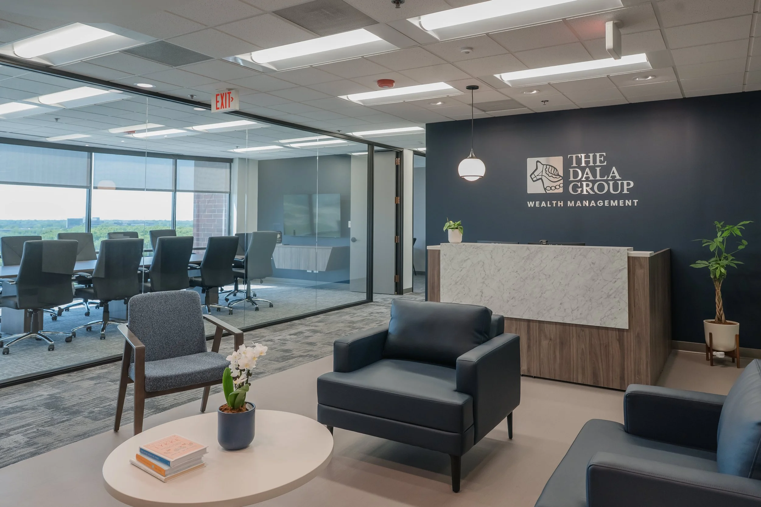 Interior view of a modern office reception area with a navy blue accent wall. The wall displays the logo and name of 'The Dala Group Wealth Management'. There is a white marble reception desk, potted plants, seating areas with chairs and sofas, a glass wall with a conference room, and large windows overlooking a cityscape and greenery.