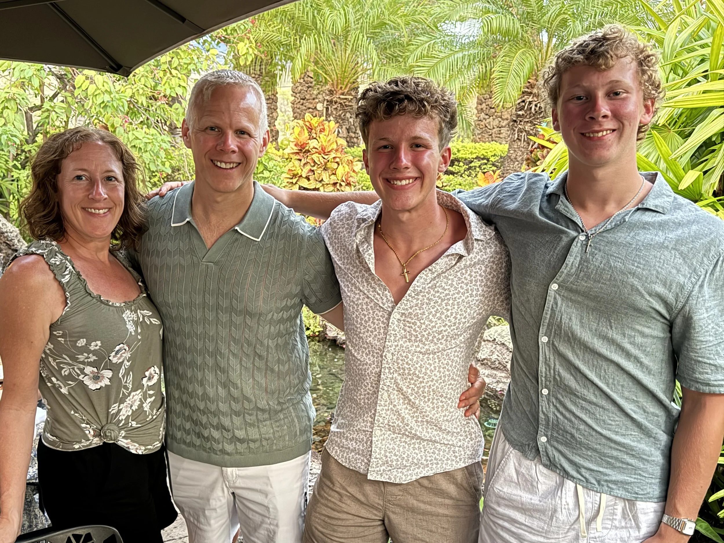 A family of five people, consisting of a woman and four young men, standing outdoors in a lush tropical garden. They are smiling and have their arms around each other.