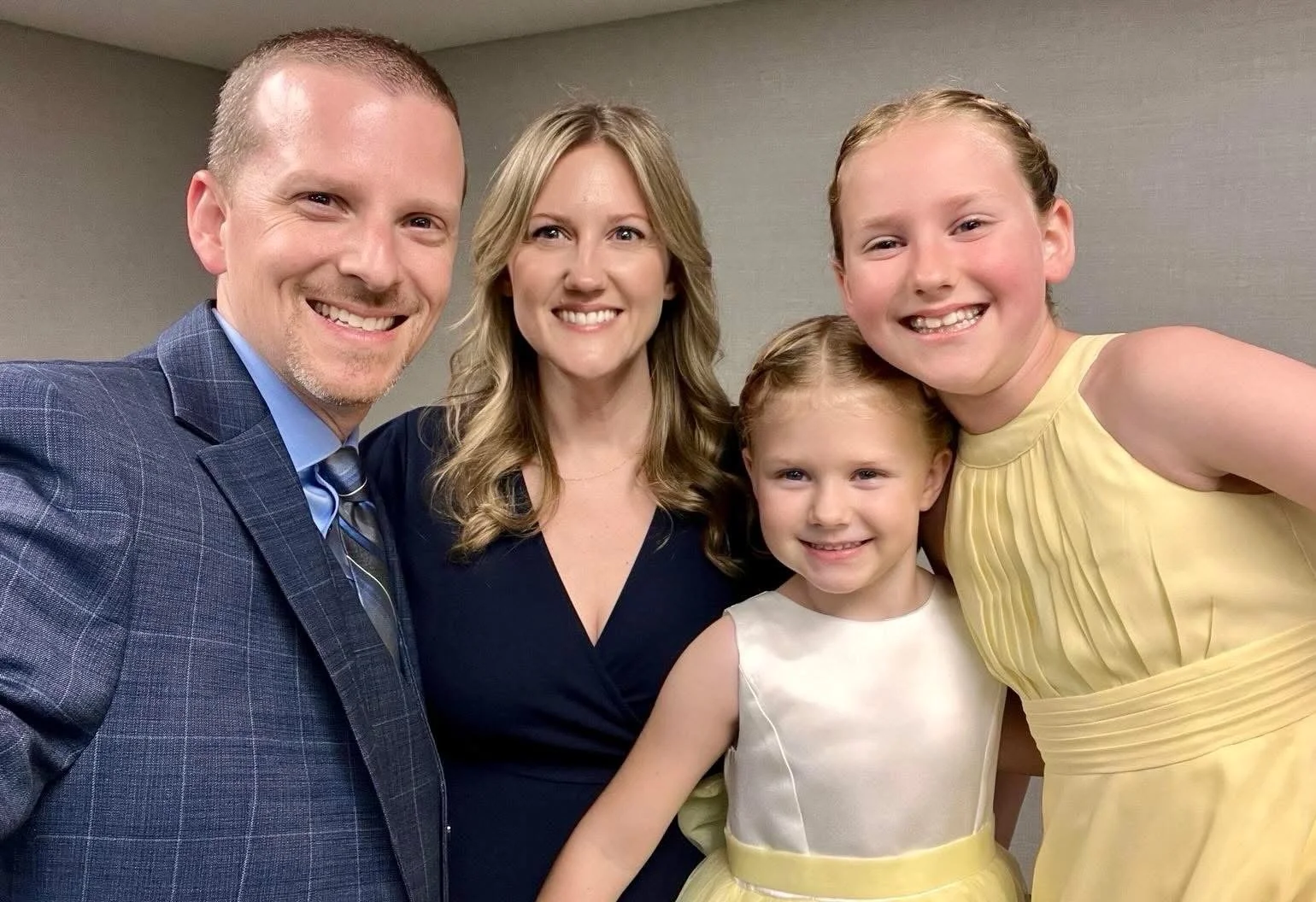 A family of five smiling for a group photo indoors, including a man in a suit, a woman in a black dress, and three young girls in pastel-colored dresses.
