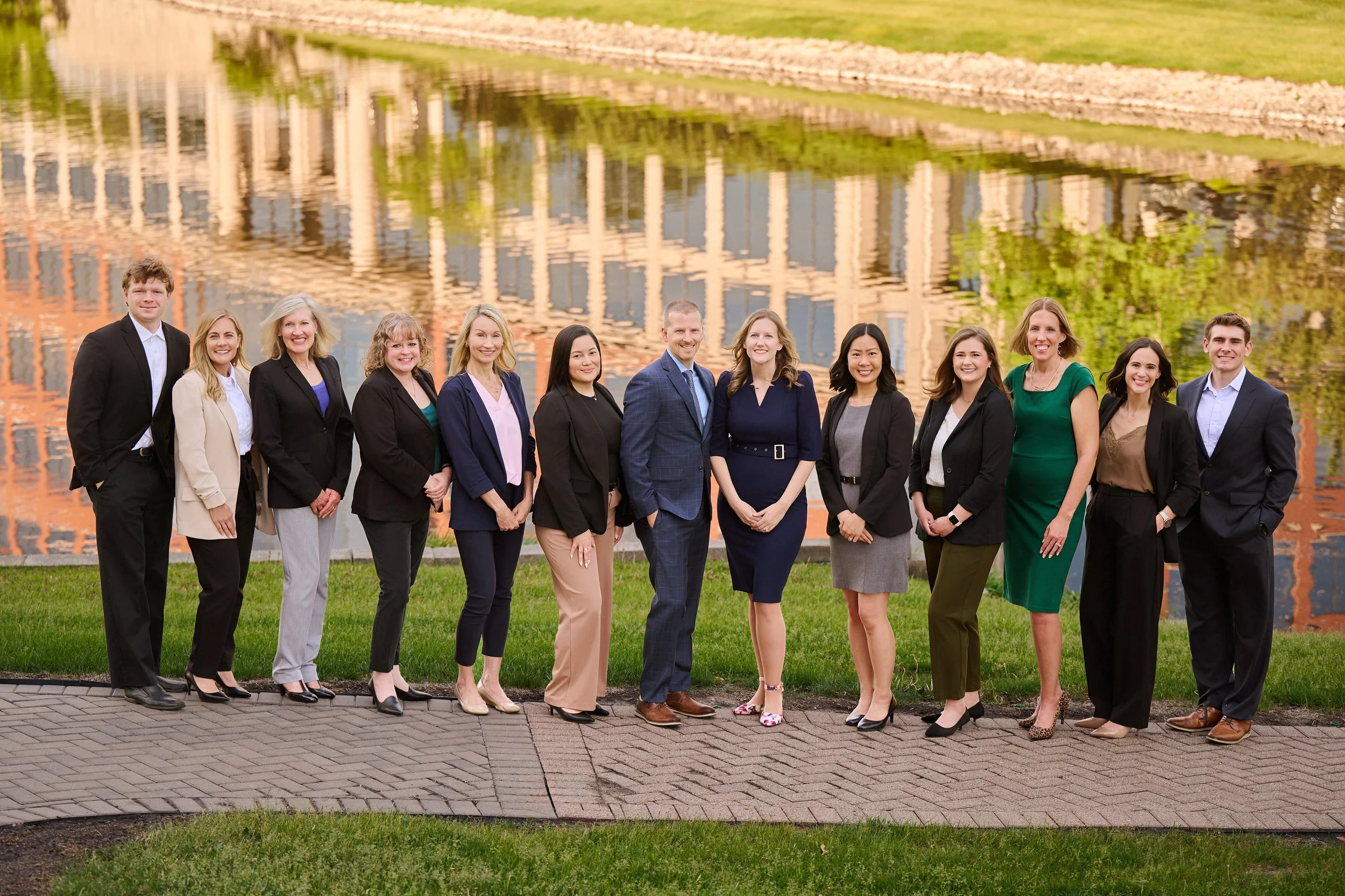 A diverse group of people in business attire standing outdoors on a brick pathway, with a grassy area and a body of water reflecting buildings in the background.