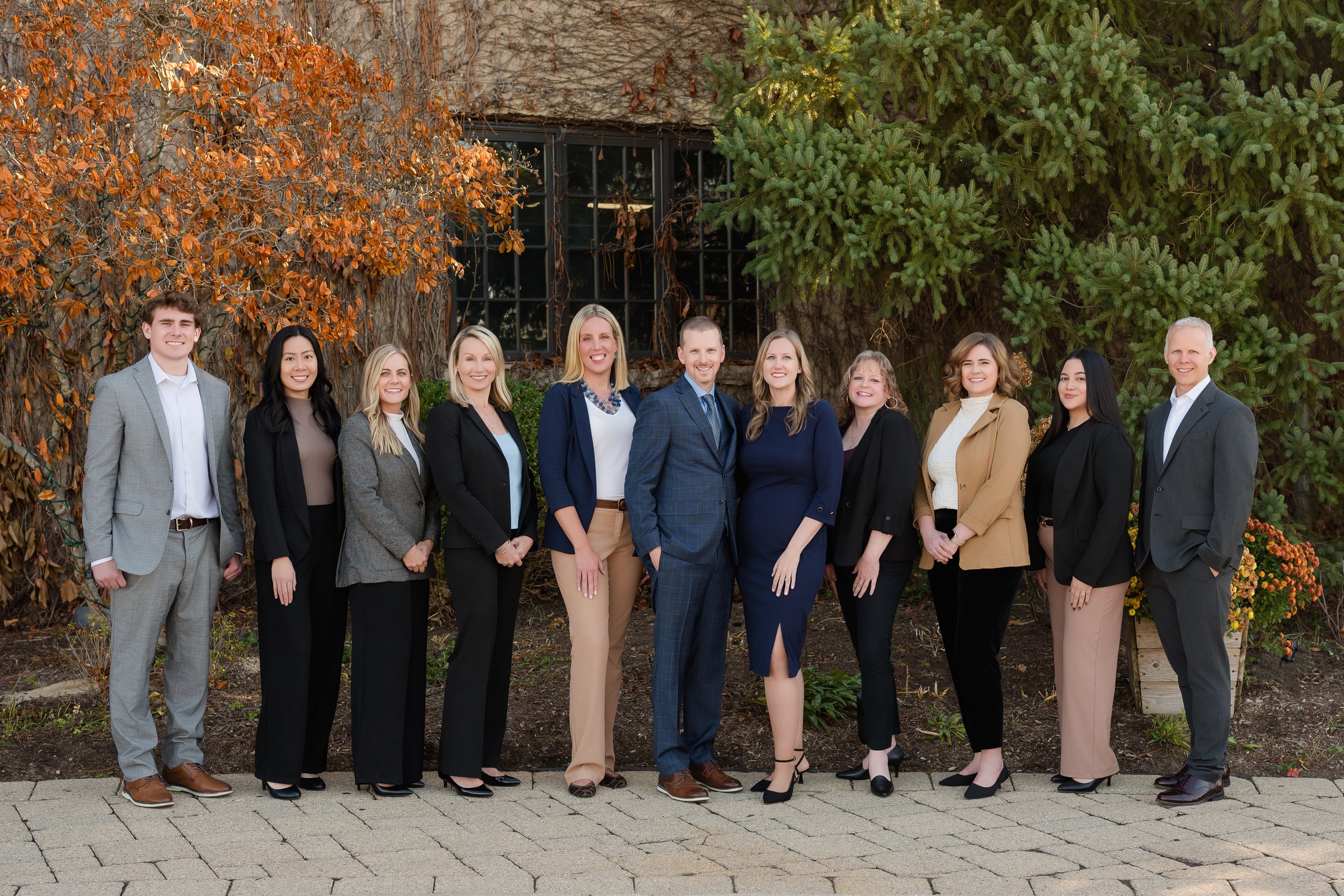 A group of twelve professionally dressed people standing outdoors on a paved pathway, smiling at the camera, with autumn leaves and trees in the background.