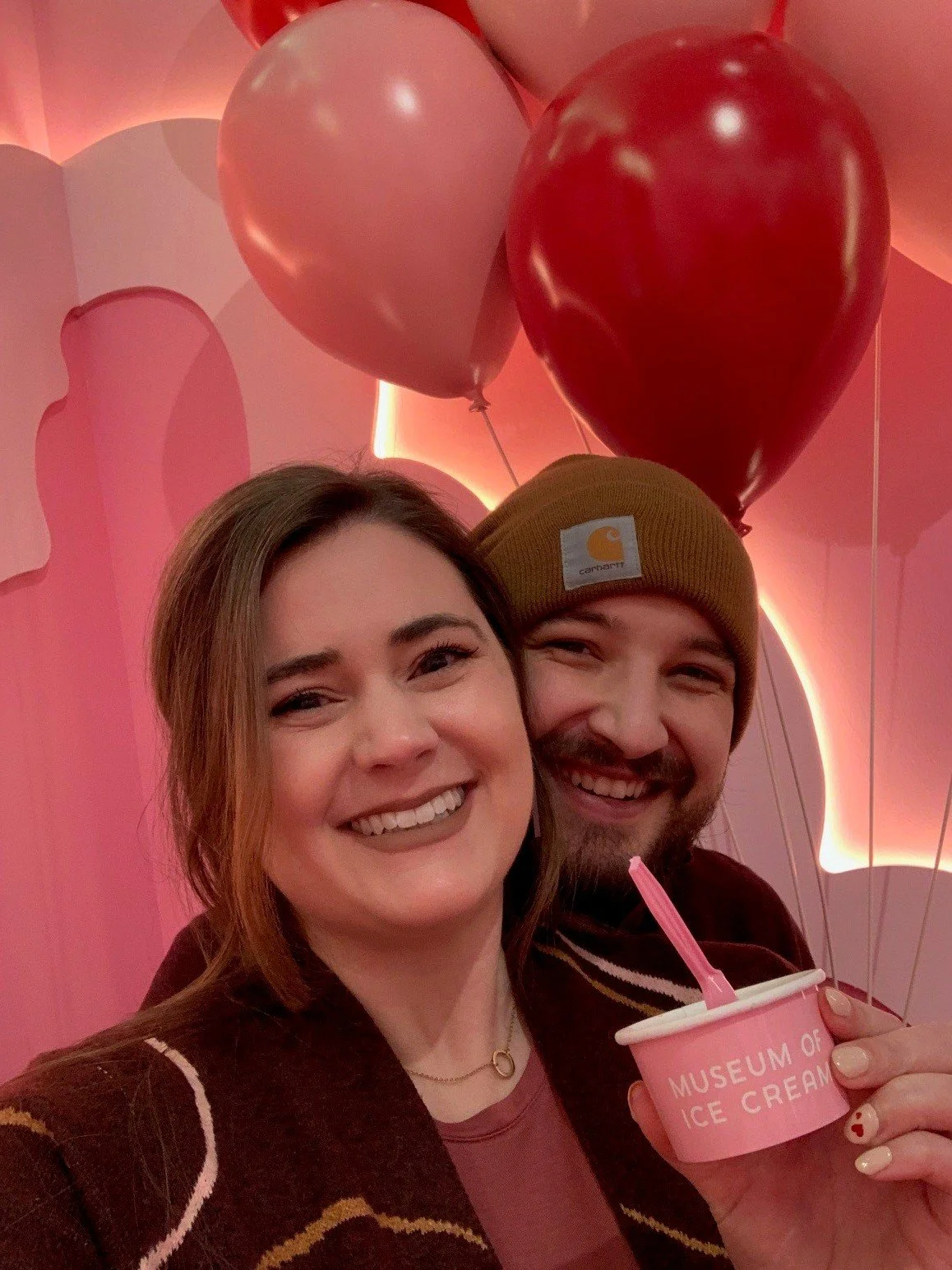 A smiling woman and man celebrating, with pink and red heart-shaped balloons, a pink container of ice cream, and a pink background with heart-shaped decorations.