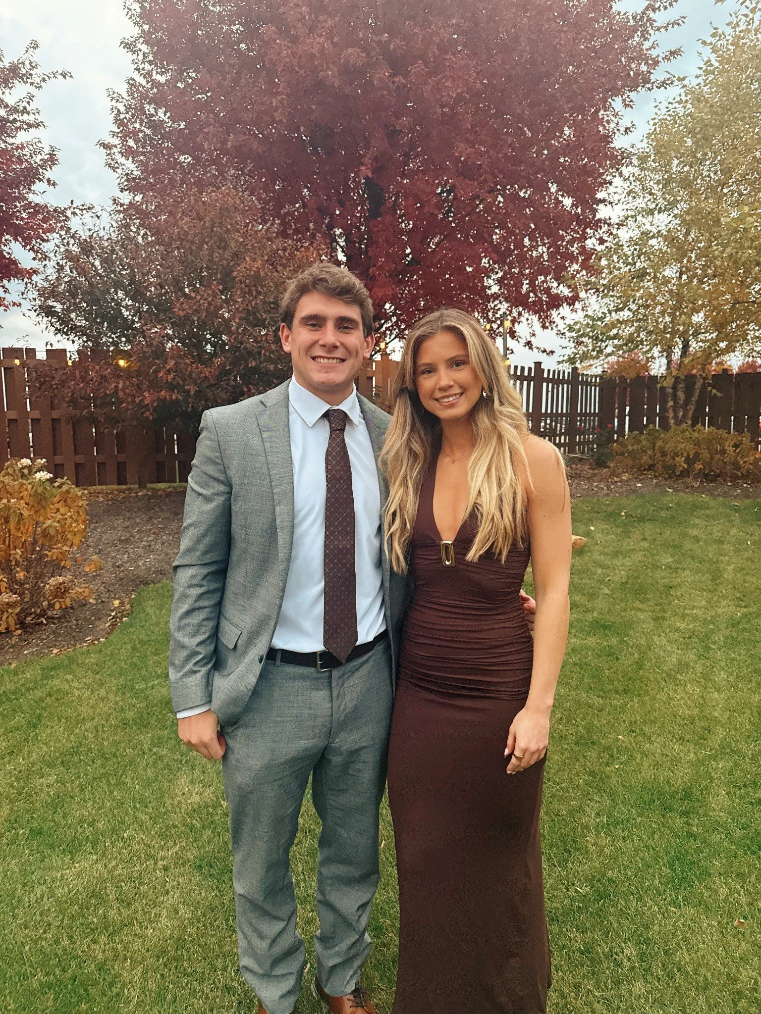 A young man and woman dressed formally, standing on a lawn with trees and a wooden fence in the background, smiling at the camera.