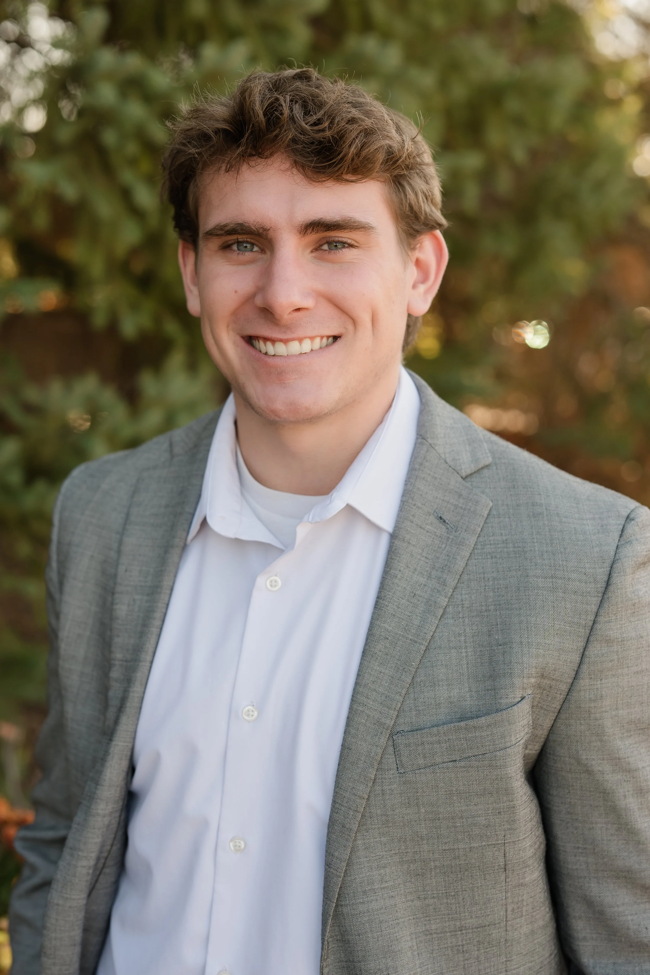 A young man with brown hair and blue eyes wearing a light gray blazer and white shirt, smiling outdoors with greenery in the background.
