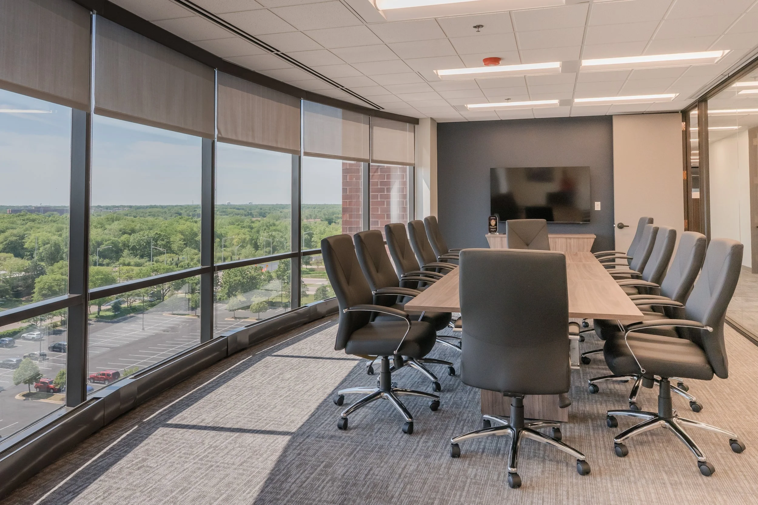 Conference room with a long wooden table surrounded by black office chairs. Large windows with blinds show a parking lot and a lush green landscape outside. A wall-mounted flat-screen TV and a small table are also visible.