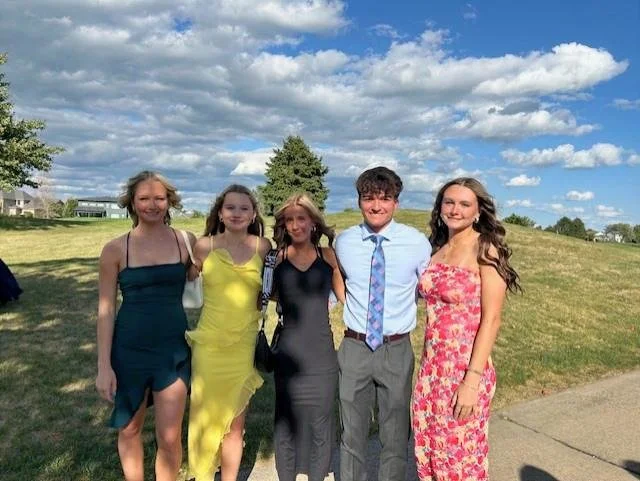 Five young people standing outdoors on a sunny day, dressed in formal and semi-formal attire, posing together on a grassy area with trees and cloudy sky in the background.