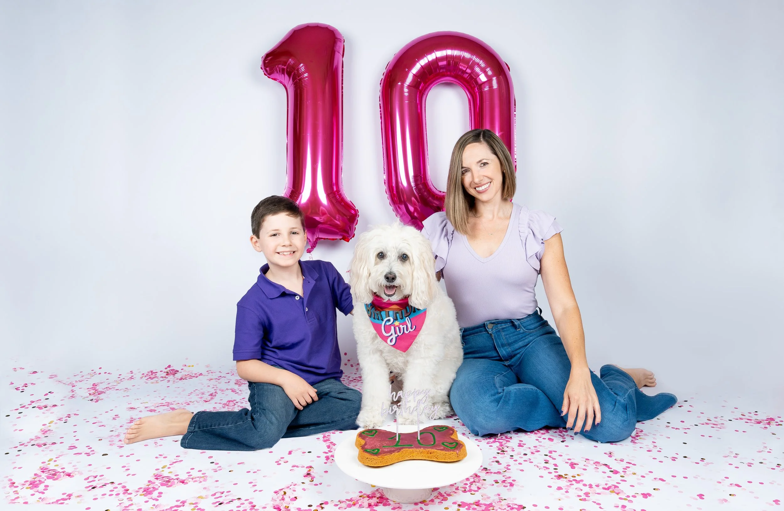 A woman, a boy, and a dog celebrating a birthday. There are pink balloons shaped as the number 10, pink confetti on the floor, and a birthday cake with a "10" candle. The woman and boy are sitting on the floor, smiling, with the woman sitting behind 