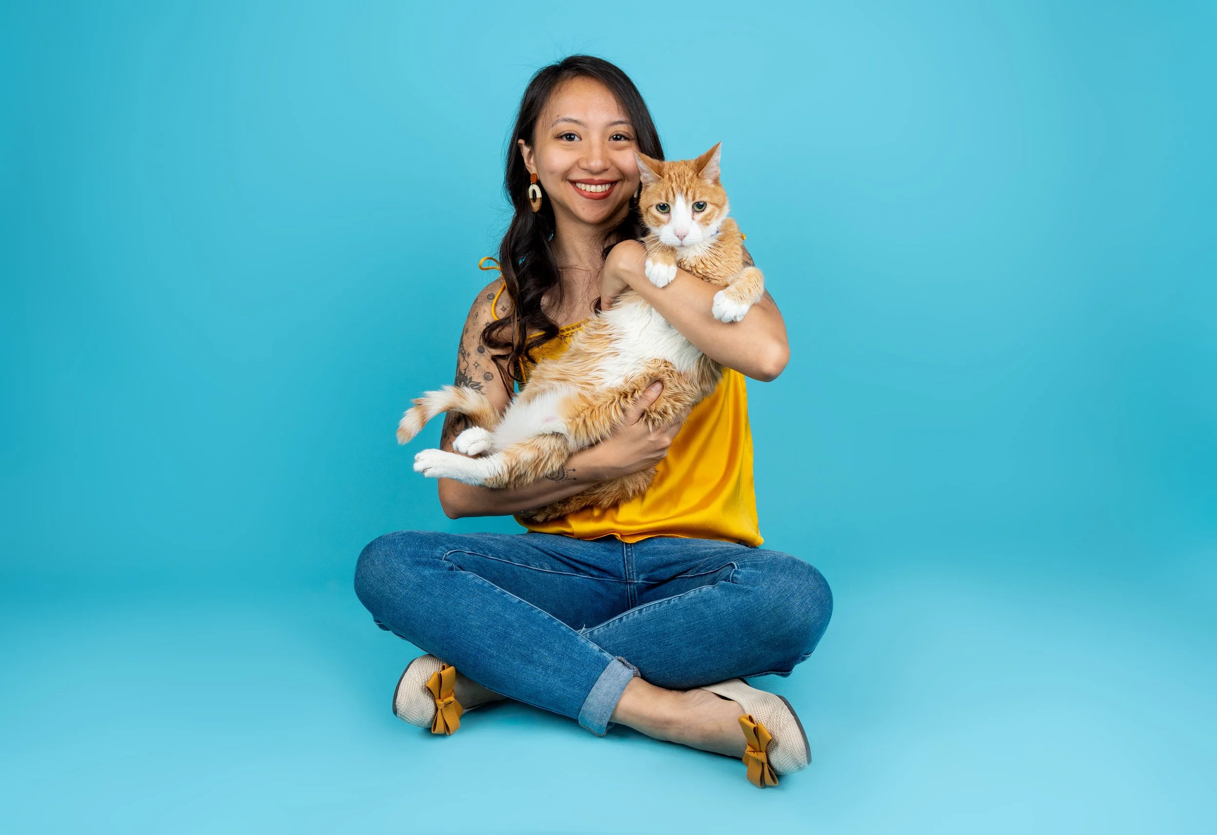 A woman with long dark hair, earrings, and a colorful tattoo on her arm, sitting cross-legged in front of a blue background, holding an orange and white cat.