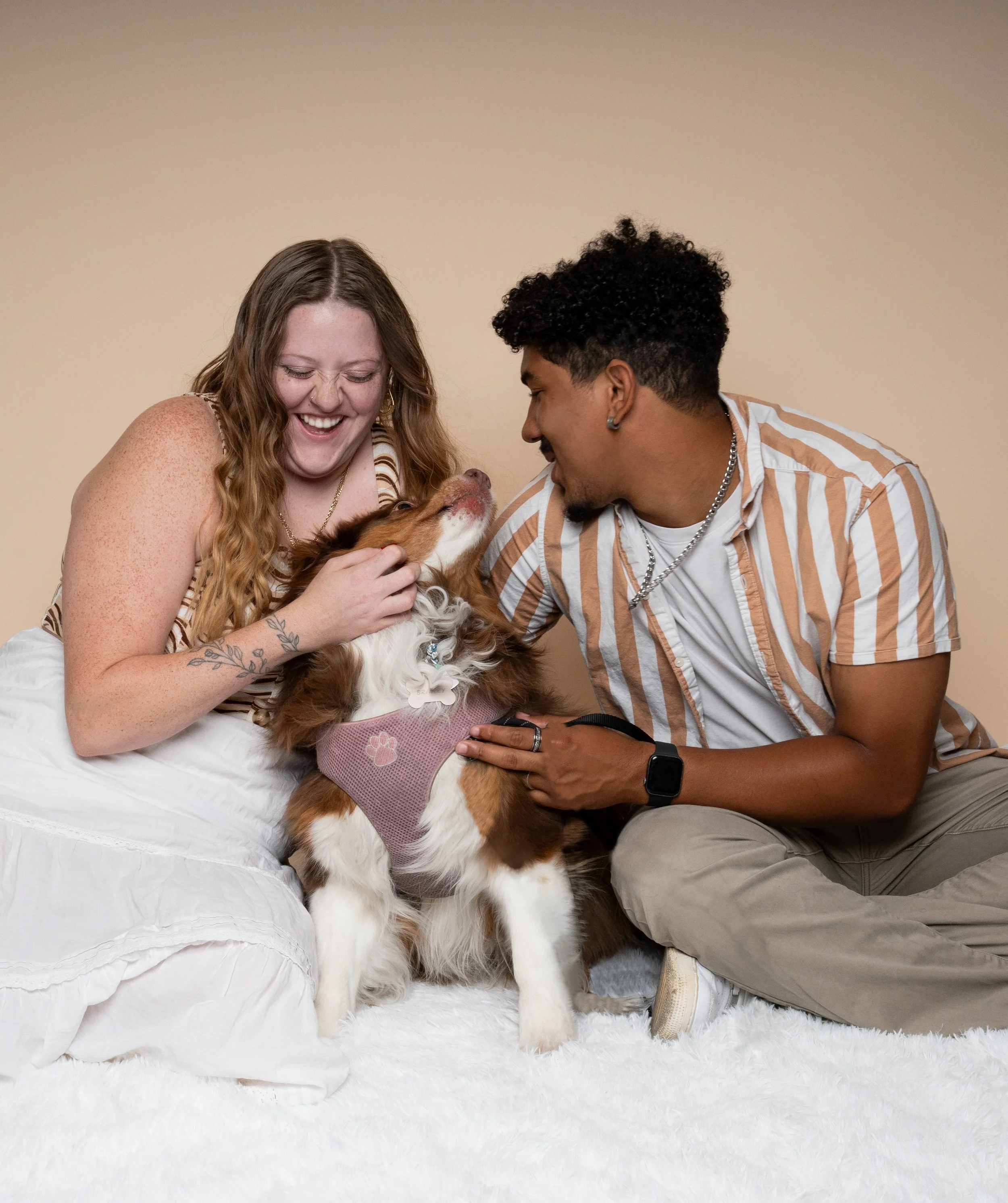 A woman with freckles and long red hair smiling and holding a Bernese Mountain Dog puppy, with a man with dark curly hair smiling at her, both sitting on a white bed against a plain beige background.