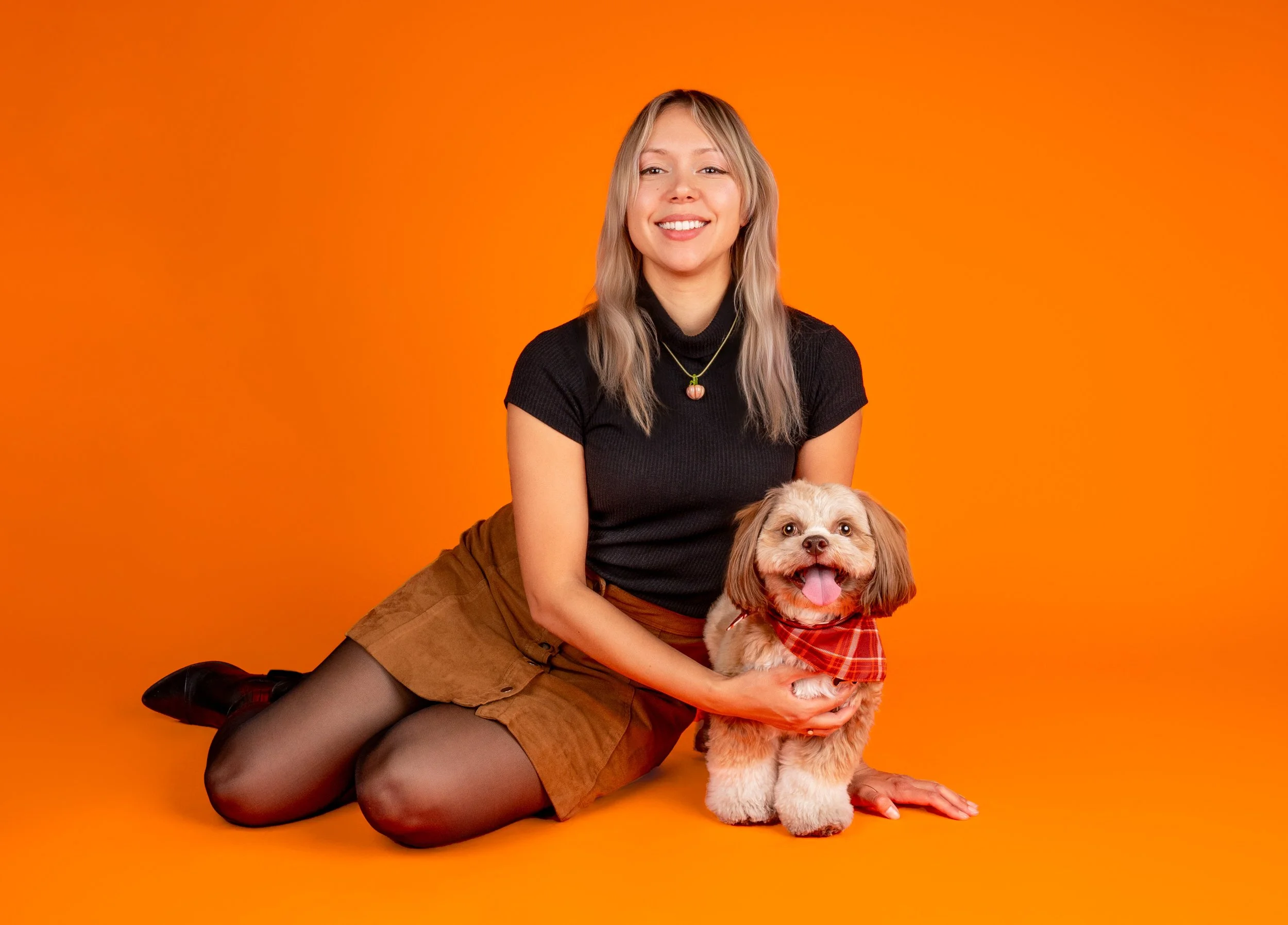 A young woman with blonde hair sitting on the floor against an orange background, holding a small brown and white dog wearing a red plaid bandana. The woman is smiling and wearing a black top, a gold necklace, and brown shorts.