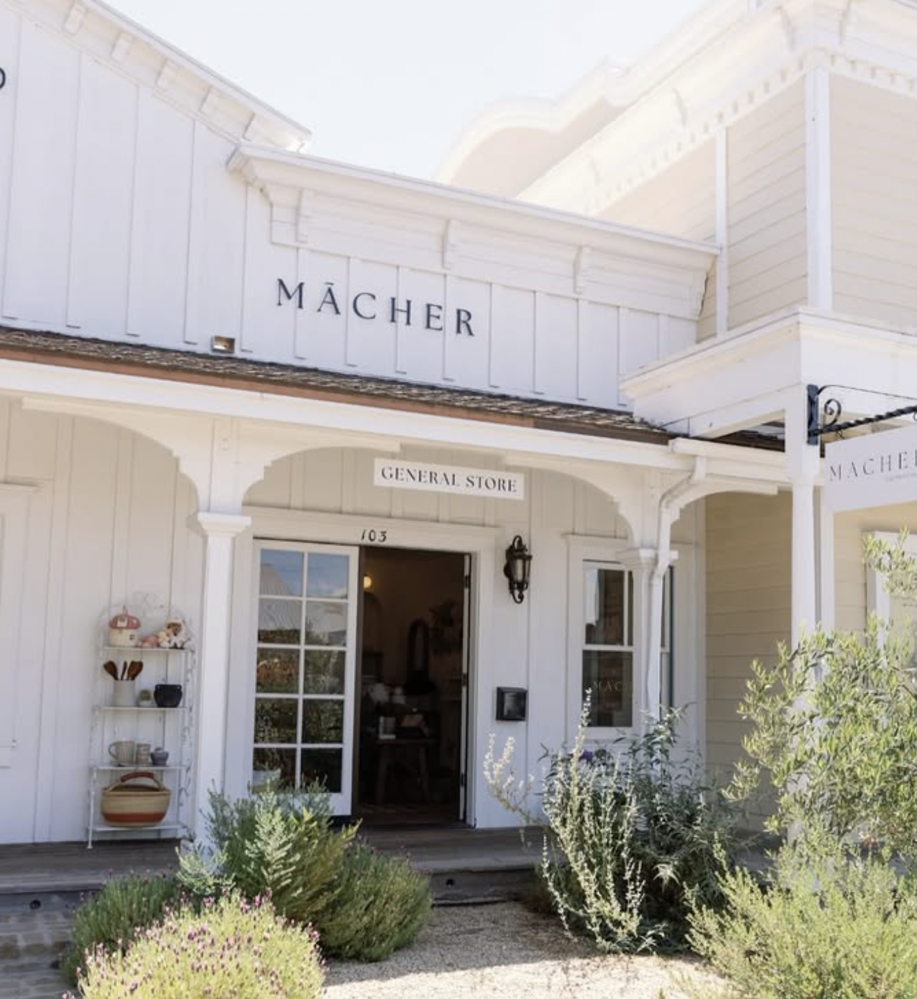 White storefront building with sign 'MÄCHER' and a smaller sign 'General Store' above the entrance, decorated with plants and shelves outside.