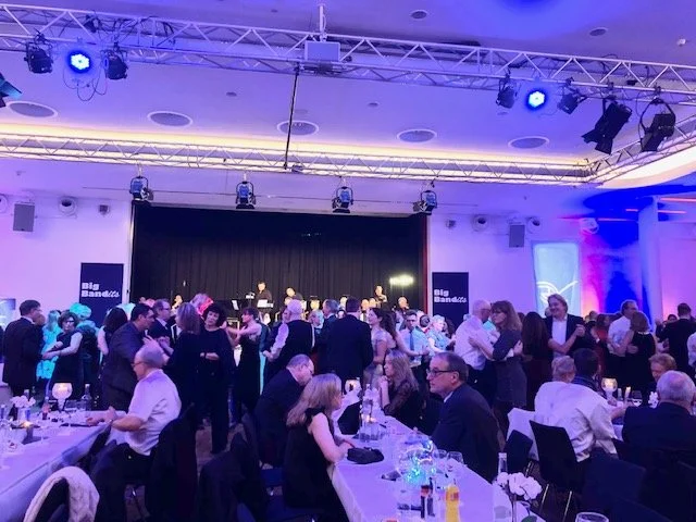 Menschen tanzen und sitzen bei einem festlichen Event im großen Saal, auf der Bühne im Hintergrund steht eine Band.