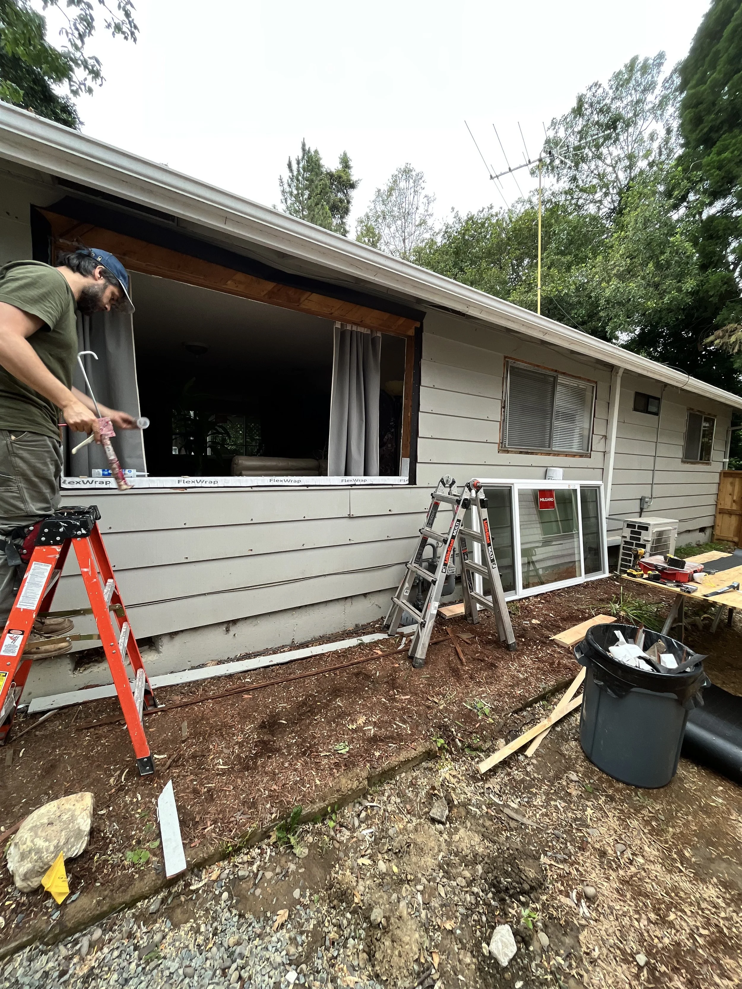 A man working on the exterior of a house, using tools and standing on a ladder. The house has a partially open window with a curtain, and construction materials and tools are scattered outside on dirt and gravel ground.