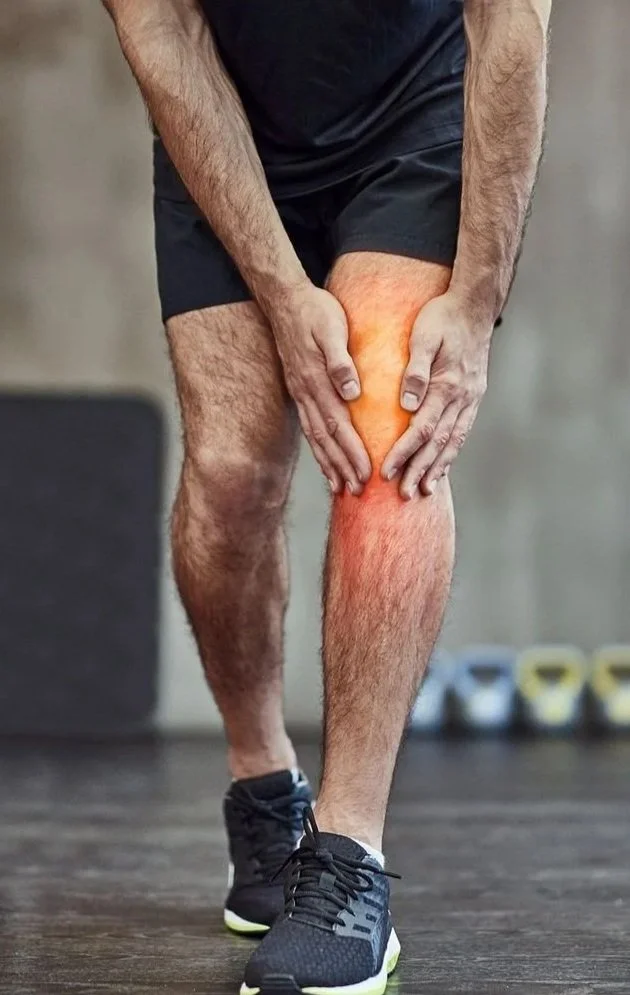 A person holding their aching or injured knee, which is highlighted in red to indicate pain or inflammation, while standing in a gym or fitness center.