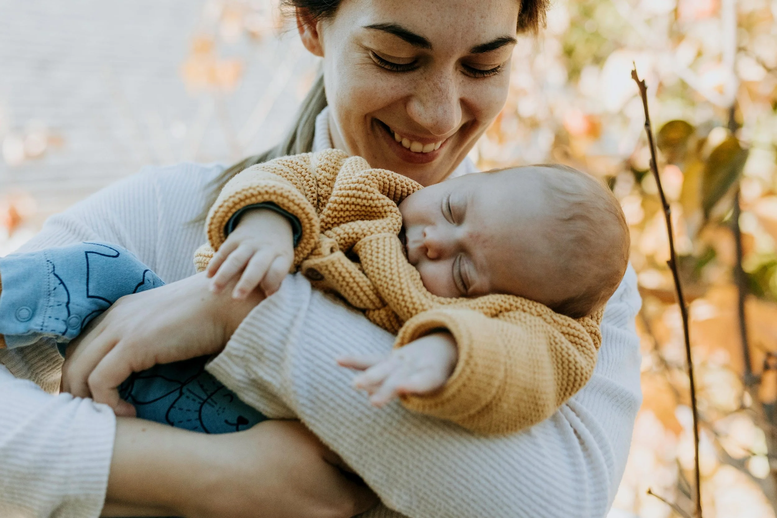 A woman holding a sleeping baby outdoors during autumn, with fall foliage in the background.