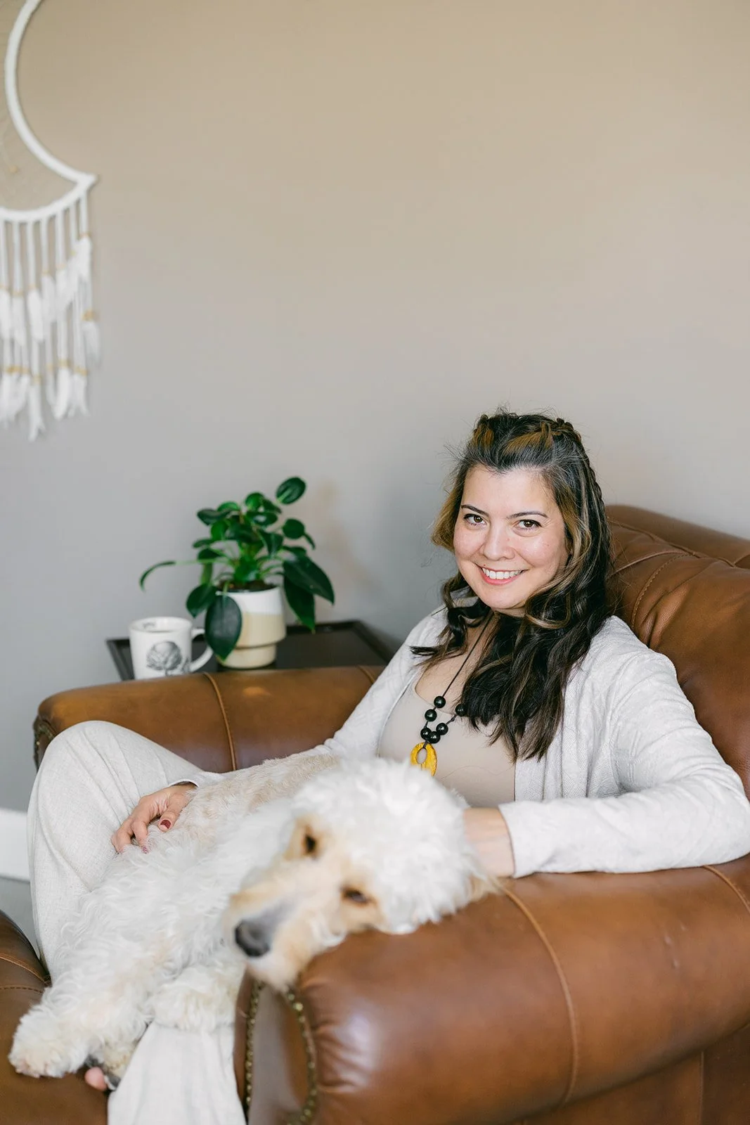 A woman with long dark hair smiling while sitting on a brown leather couch, with a white fluffy dog lying beside her, in a cozy living room with a plant and a mug on a side table.