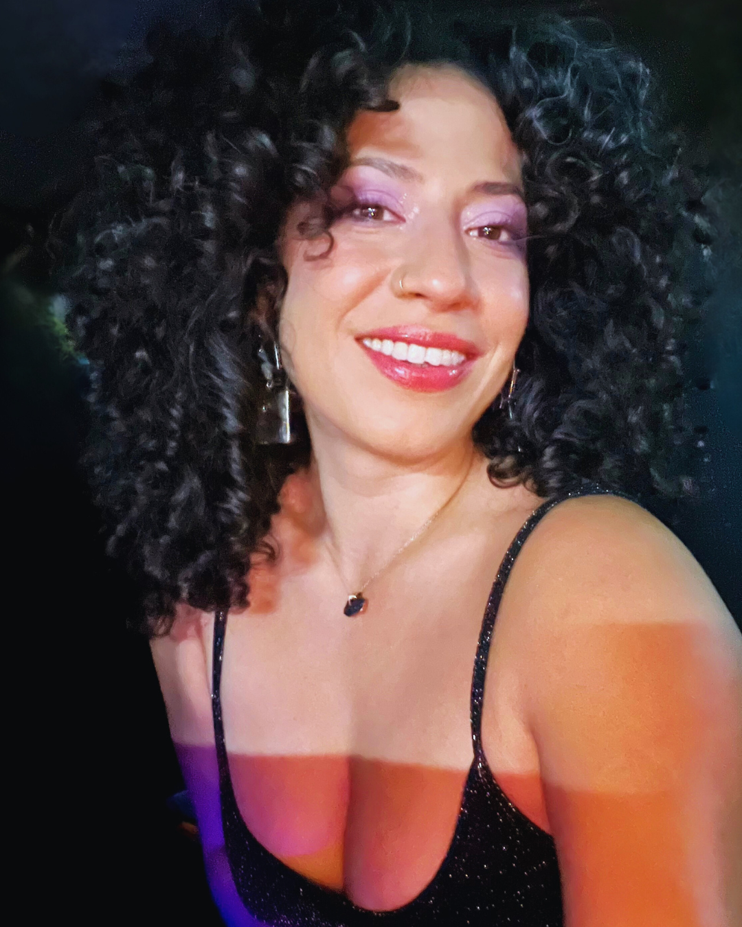 A woman with dark curly hair smiling at the camera, wearing a black sparkly dress with thin straps, pink eyeshadow, and jewelry including earrings, a necklace, and a nose ring.
