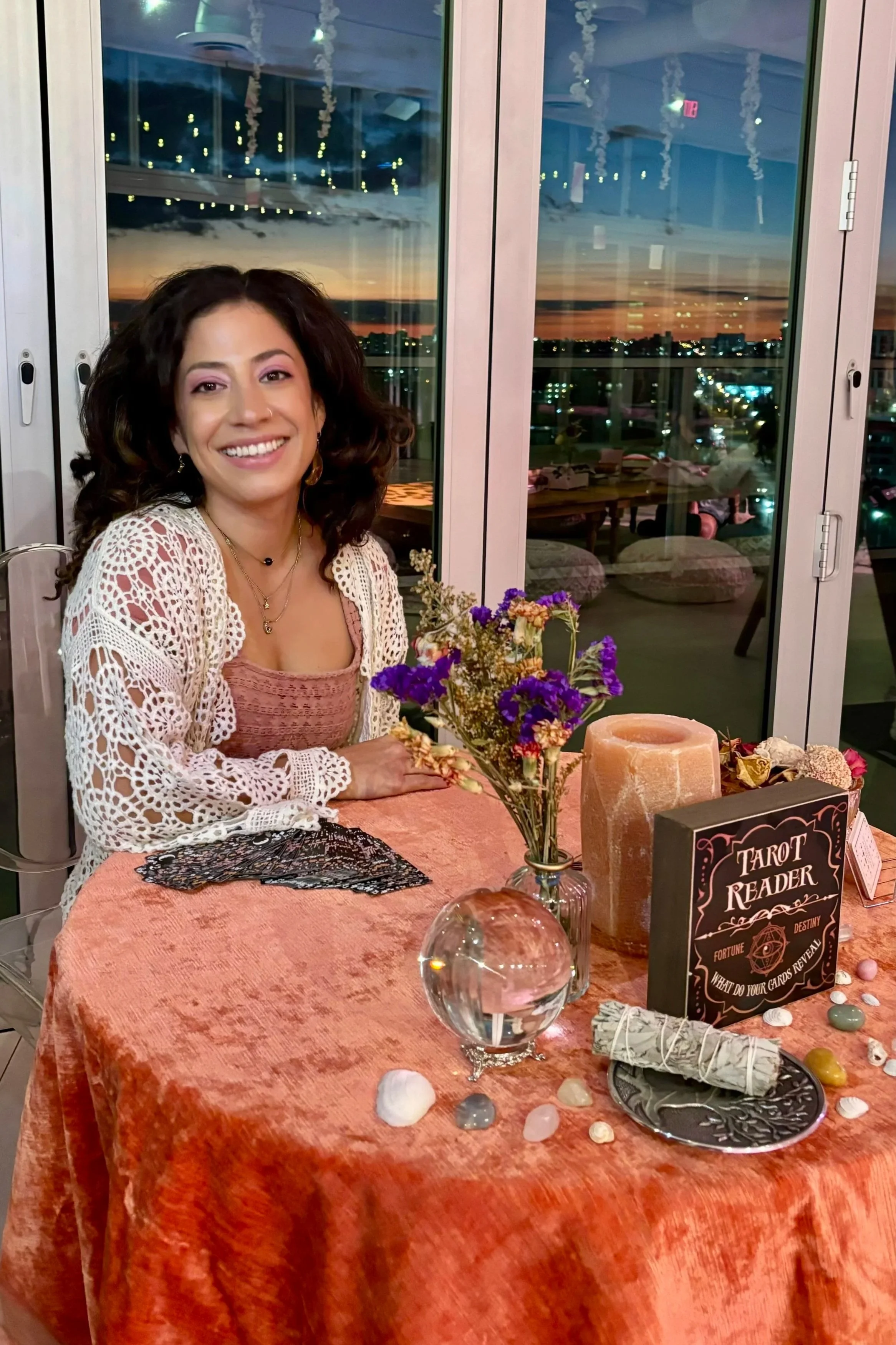 A woman with dark, curly hair sitting at a table with tarot reading items, including crystal, shells, and a sign reading 'Tarot Reader'. She is smiling and wearing a crochet white top over a pinkish dress, with a sunset view through glass doors behind her.