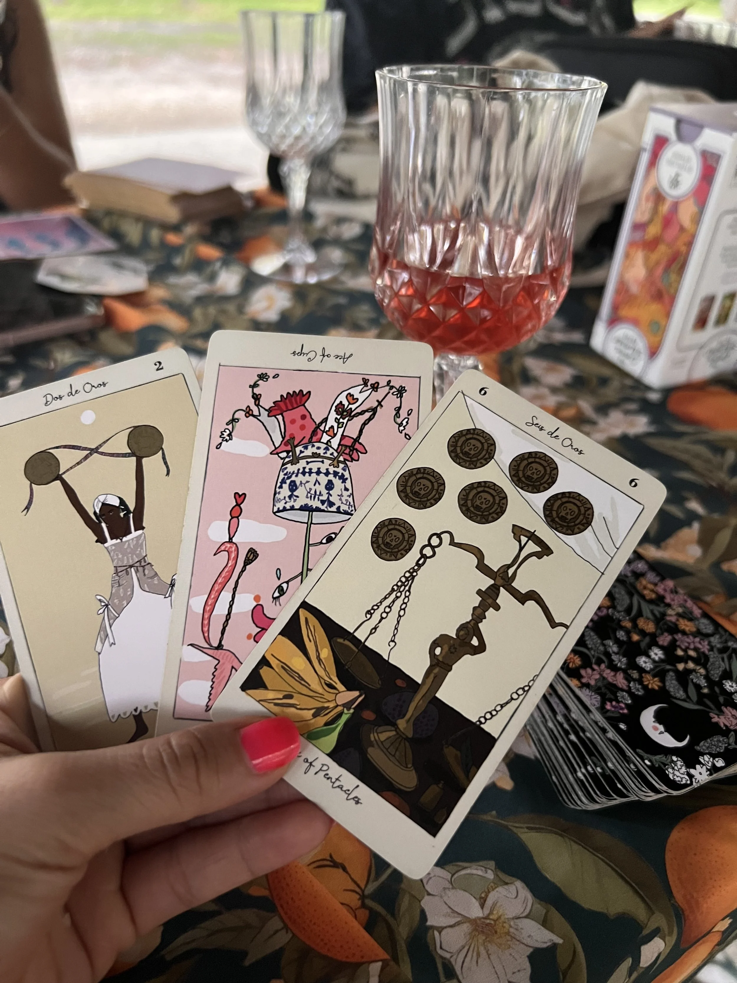 A hand holding three tarot cards over a table with a glass of pink wine, a wine glass, a box of tarot cards, and a notebook in the background.