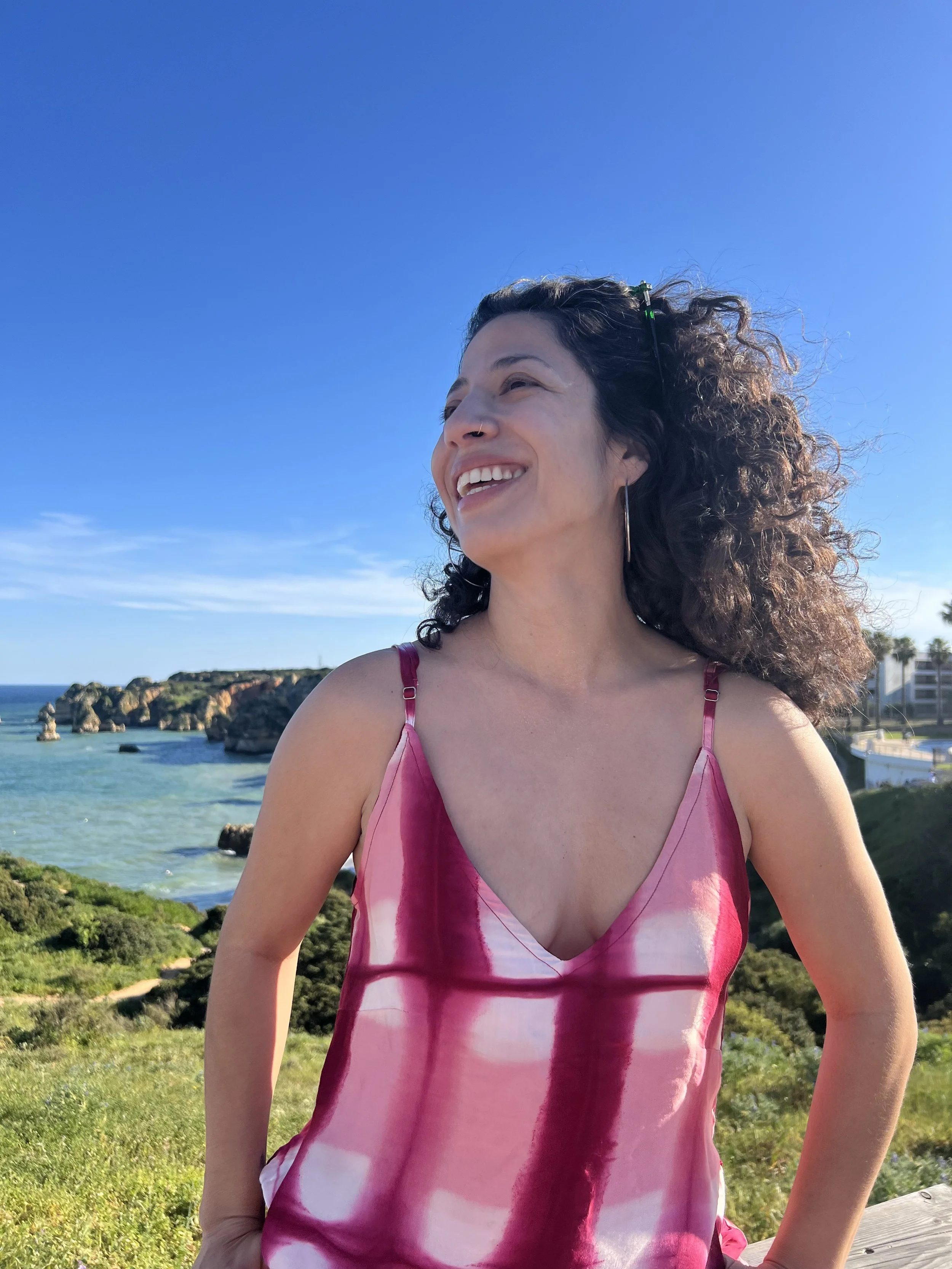 A woman with curly hair smiling in a pink and white dress at a scenic coastal location with rocks, ocean, and greenery in the background on a sunny day.