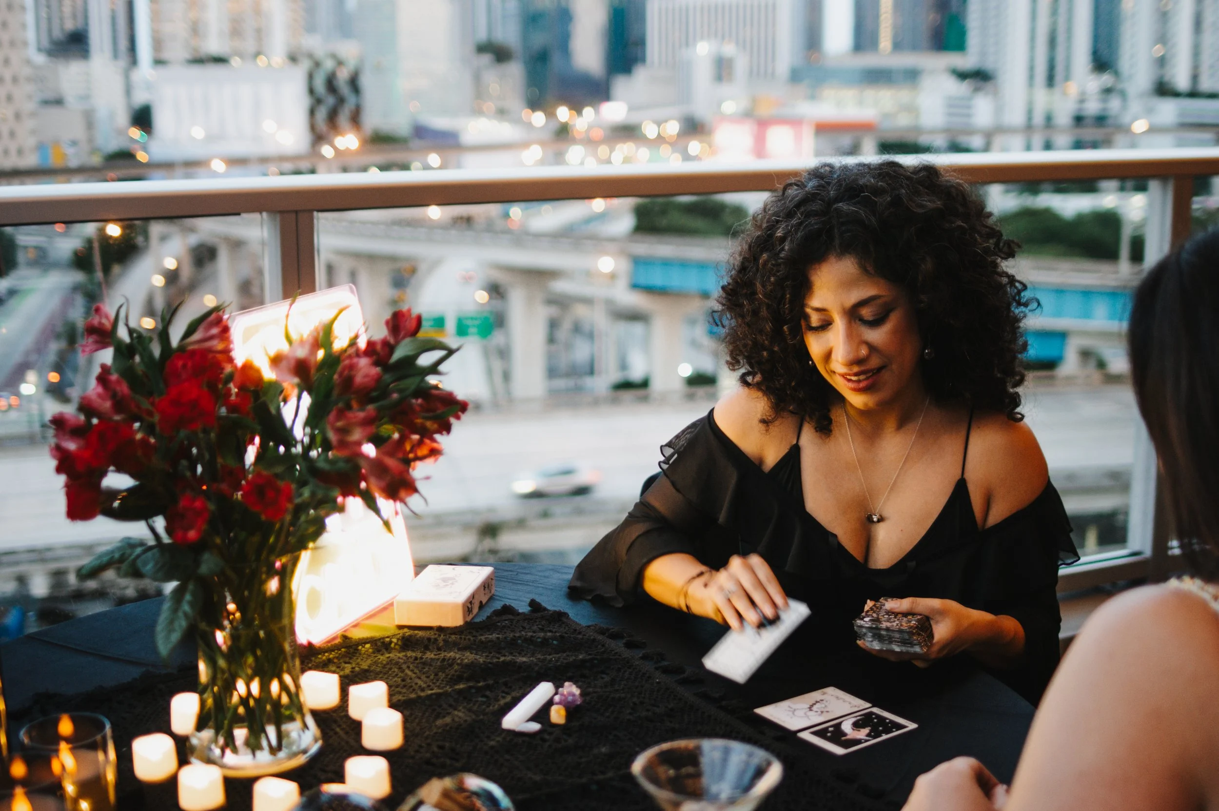 A woman with dark curly hair wearing a black off-the-shoulder top, sitting at a table with tarot cards and crystals, appearing to be conducting a tarot reading. The table is decorated with a vase of red flowers, small candles, and a cozy ambient lighting.