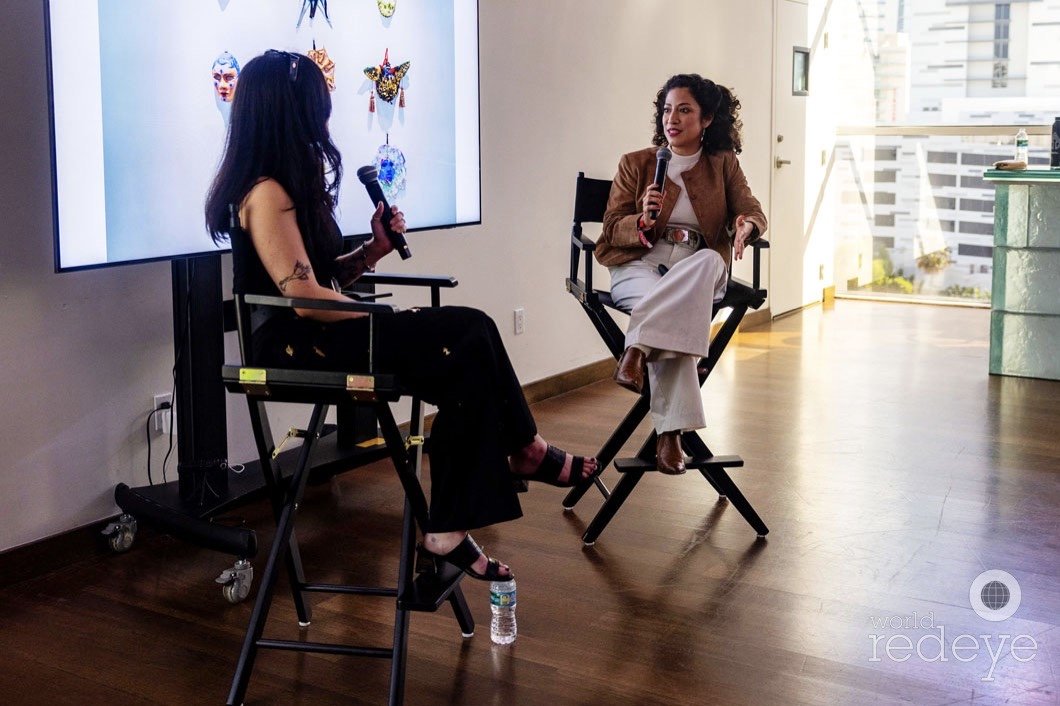 Two women are sitting on director's chairs, engaging in a discussion with one holding a microphone and a water bottle on the floor nearby. A large screen behind them displays colorful masks and face art. The setting appears to be a bright room with wooden floors and floor-to-ceiling windows.
