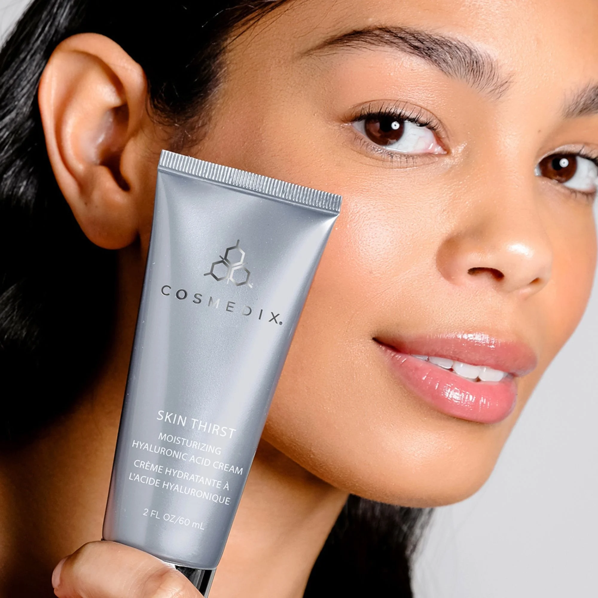 Woman holding a silver tube of COSMEDIX Skin Thirst hyaluronic acid moisturizer beside her face.