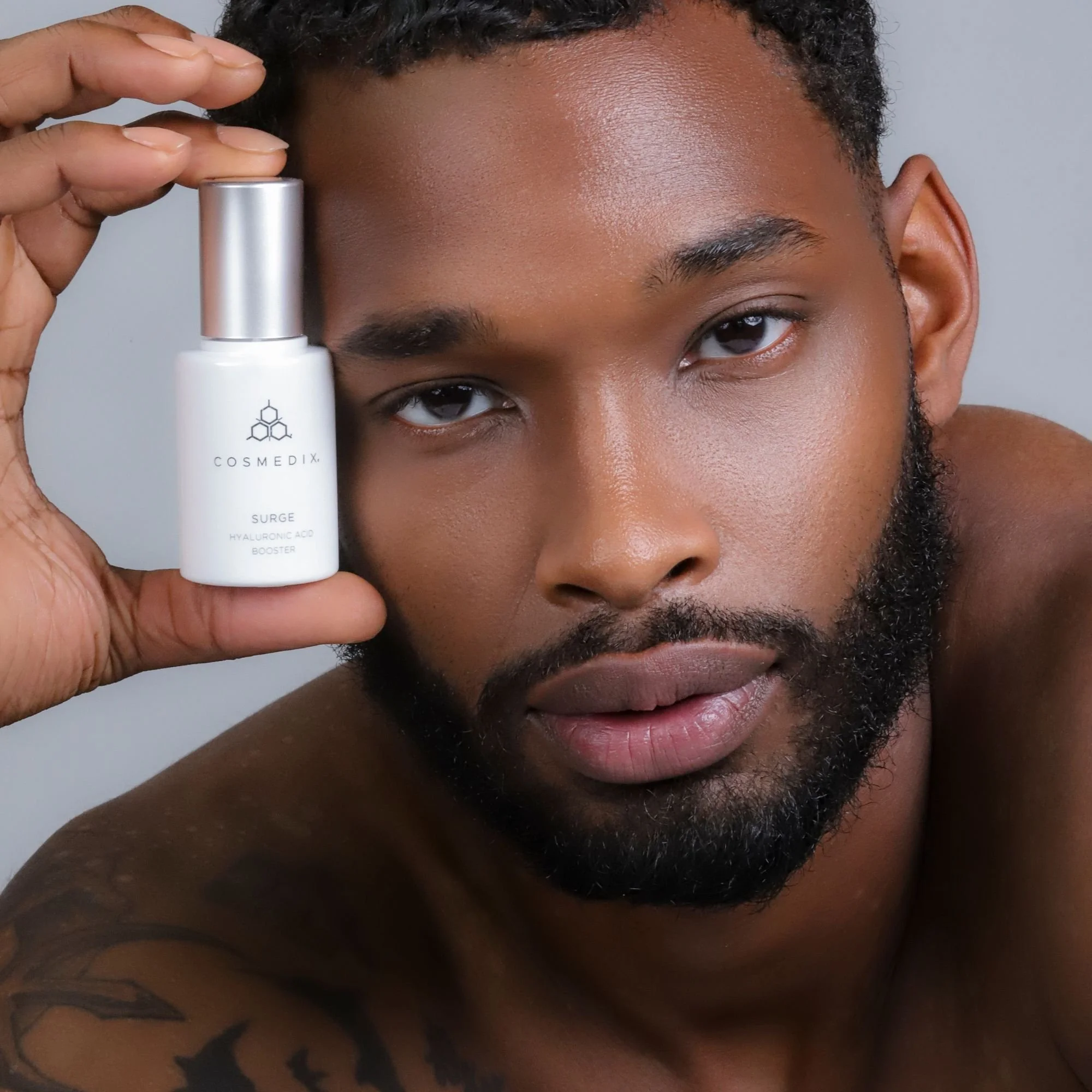 Man holding a bottle of COSMEDIX Surge Hyaluronic Acid Booster beside his face against a gray background.