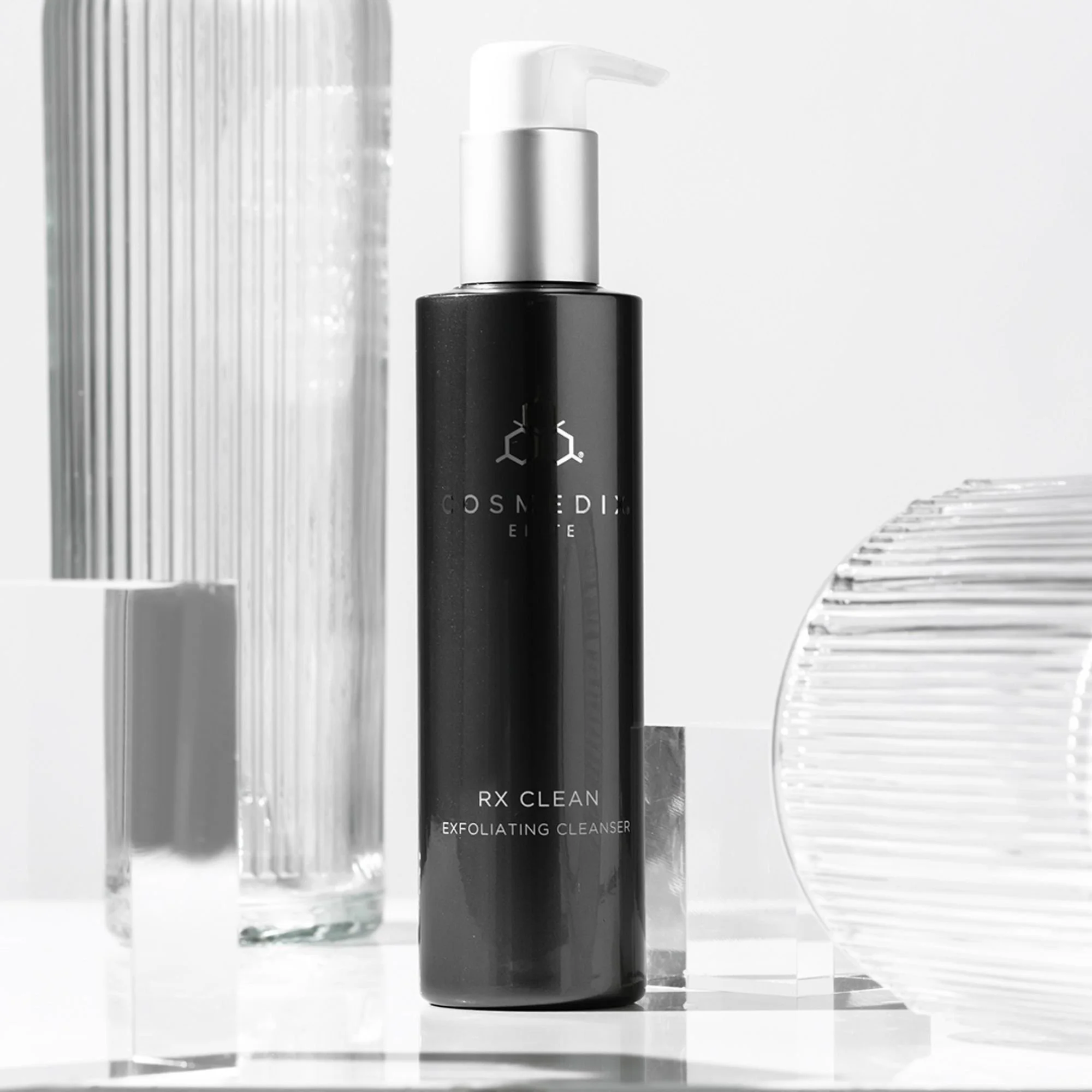 COSMEDIX Elite RX Clean Exfoliating Cleanser black pump bottle displayed beside clear ribbed glass and acrylic objects.