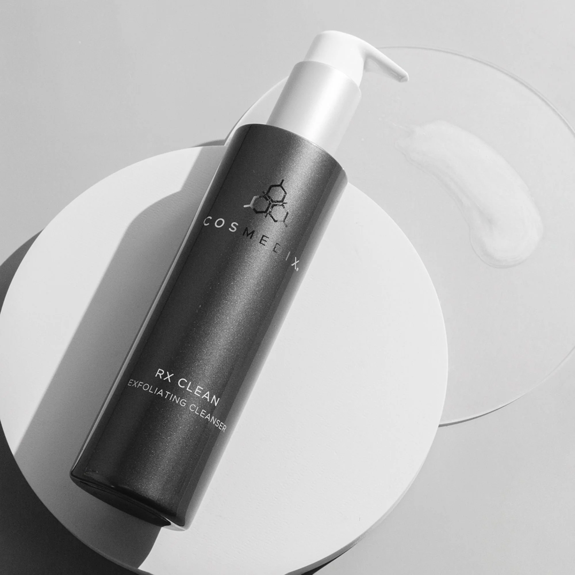 COSMEDIX Elite RX Clean Exfoliating Cleanser bottle positioned diagonally on a round white display surface.