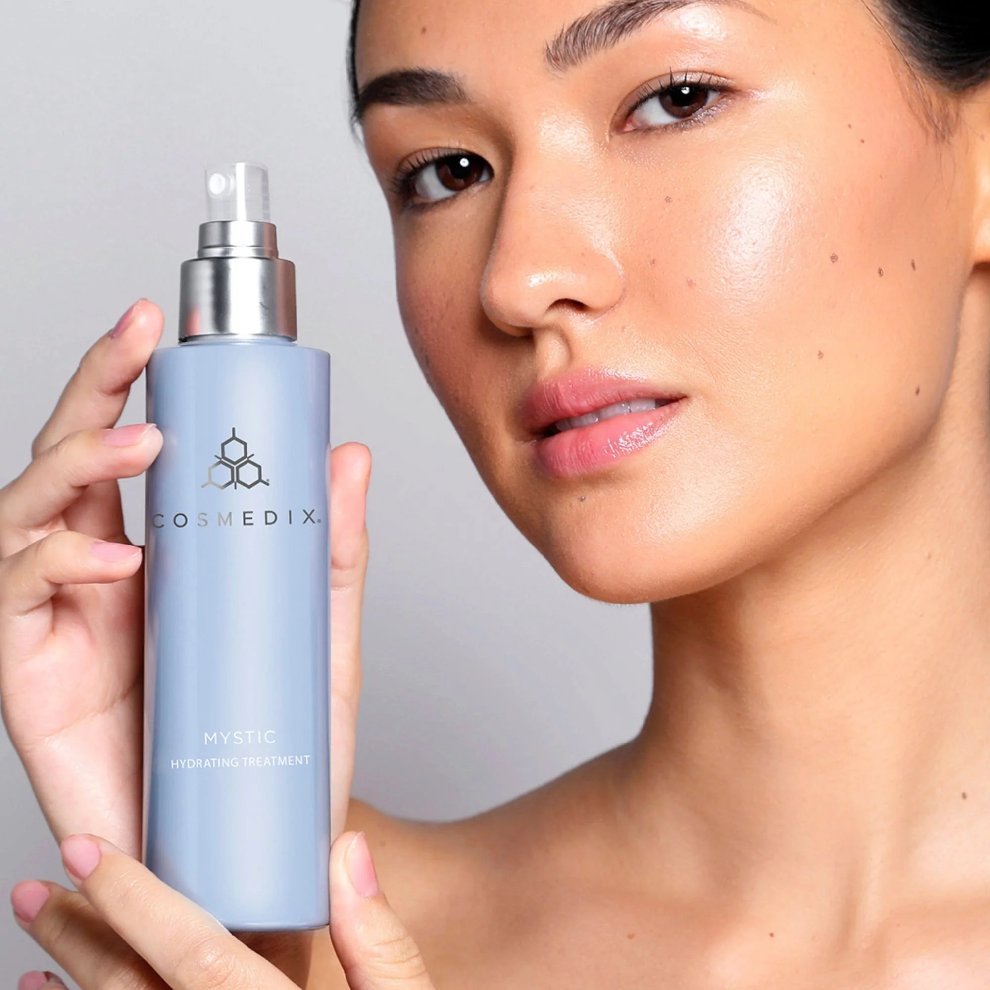 Model holding a COSMEDIX Mystic Hydrating Treatment bottle beside her face.