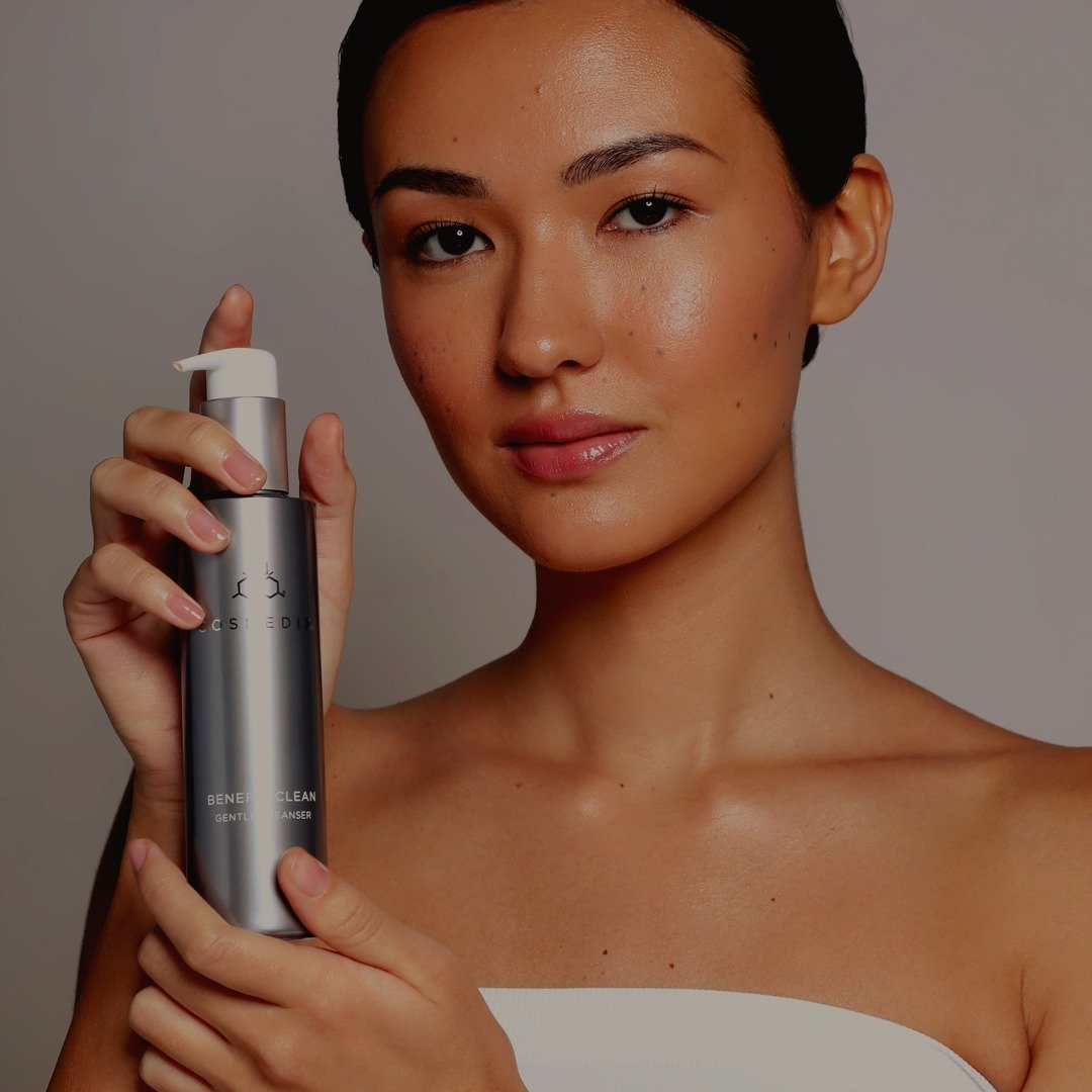 Woman holding a COSMEDIX Benefit Clean Gentle Cleanser bottle beside her face in a skincare product portrait.