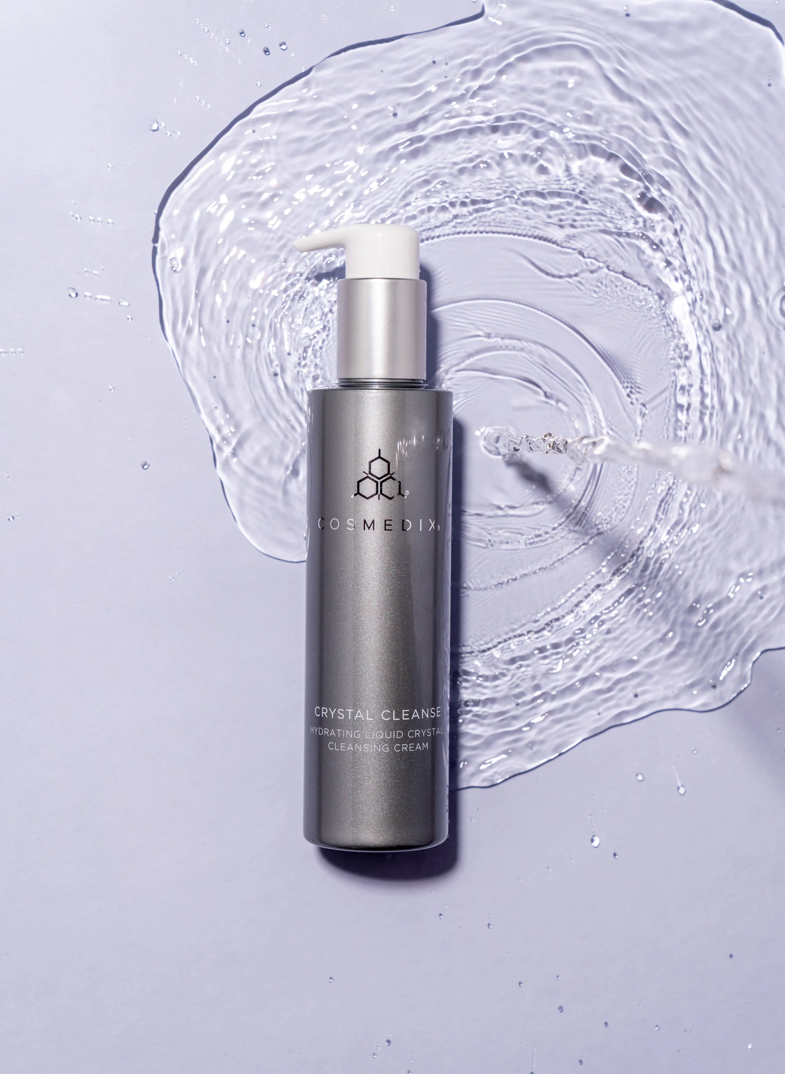 COSMEDIX Crystal Cleanse silver pump bottle positioned diagonally beside a white cream swatch on a pale lavender background.