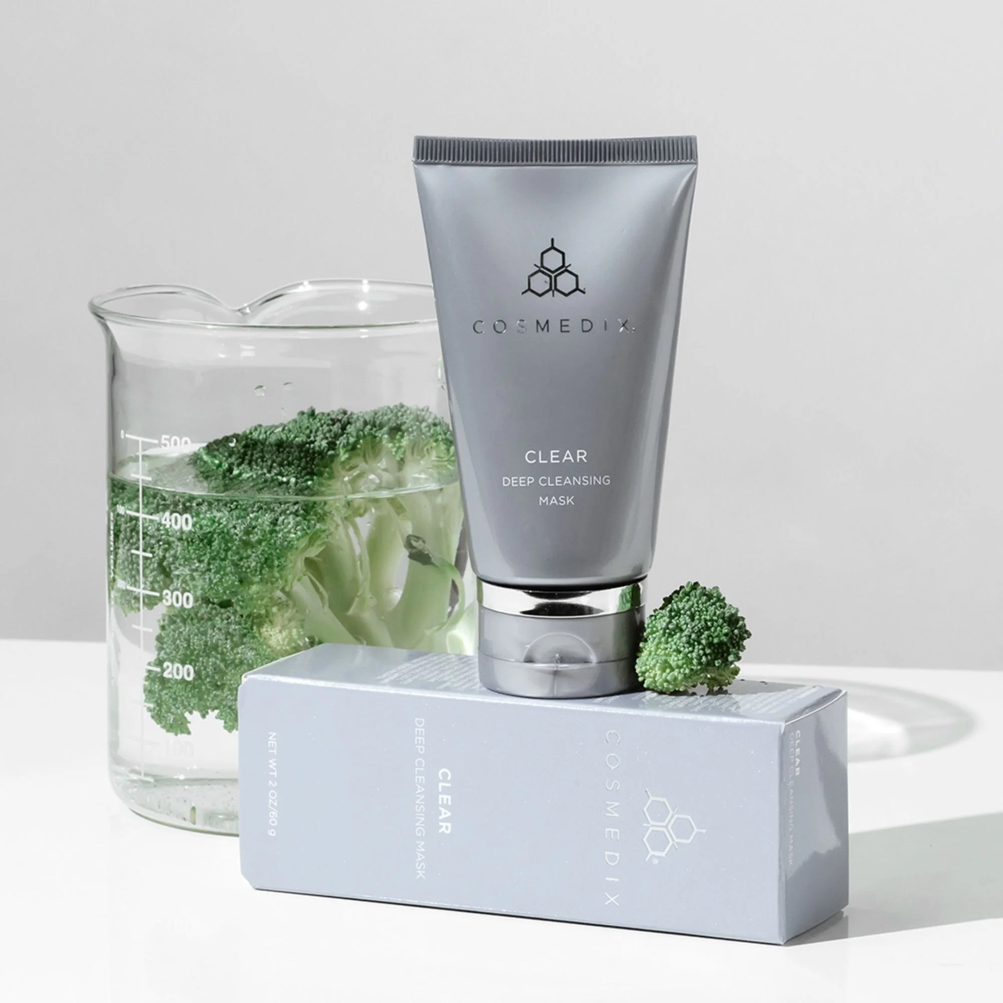 COSMEDIX Clear mask tube standing on its box beside broccoli and a glass beaker against a light gray background.