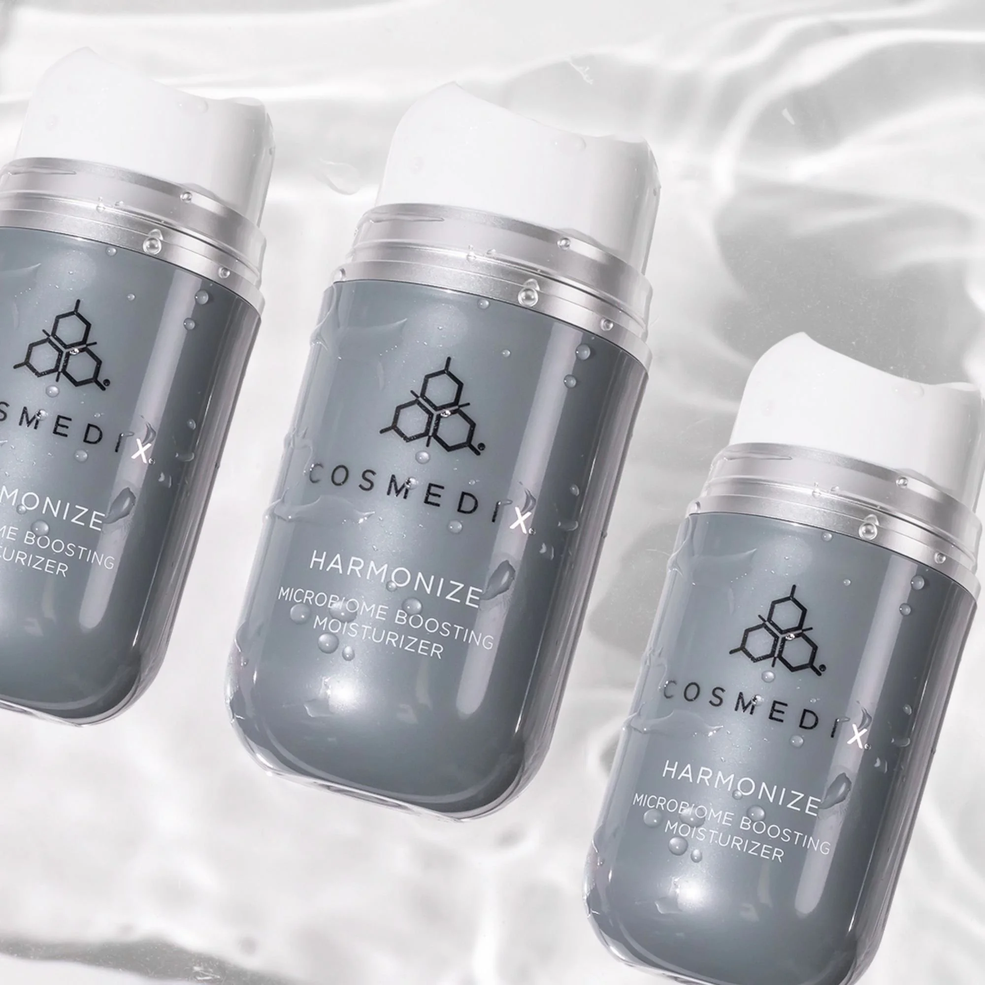 Three COSMEDIX Harmonize moisturizer bottles covered in water droplets against a rippling water background.