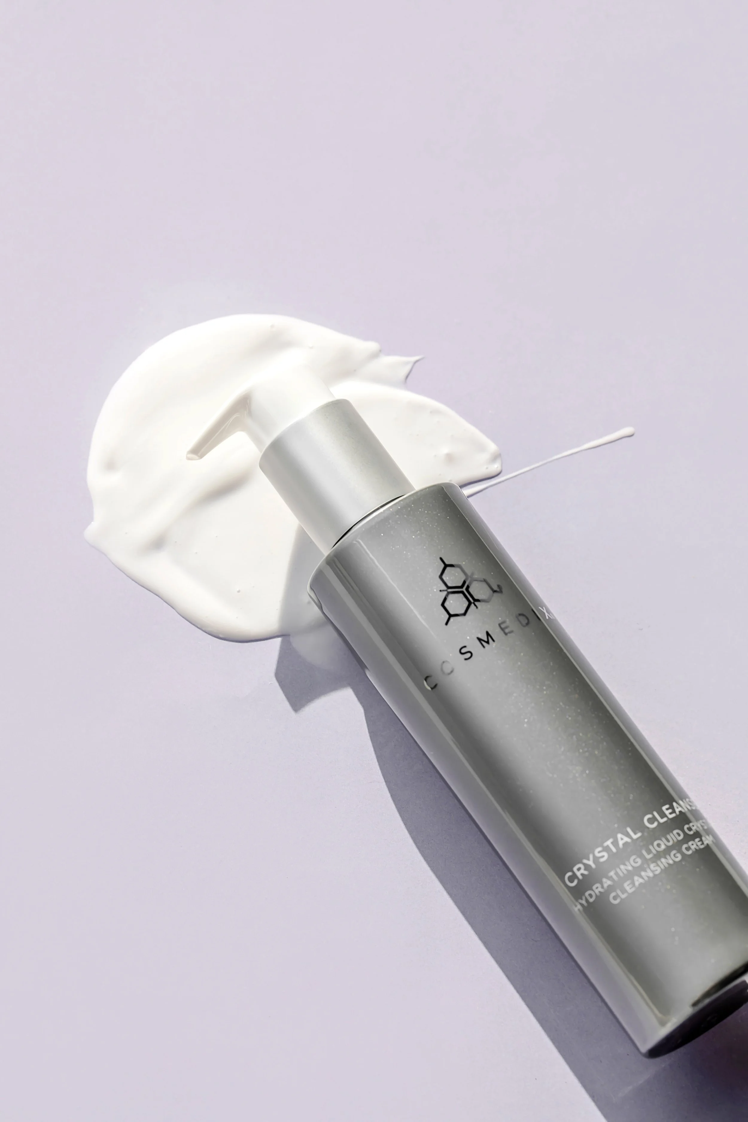COSMEDIX Crystal Cleanse silver pump bottle displayed diagonally on a light stone surface beside drops of white cleansing cream.