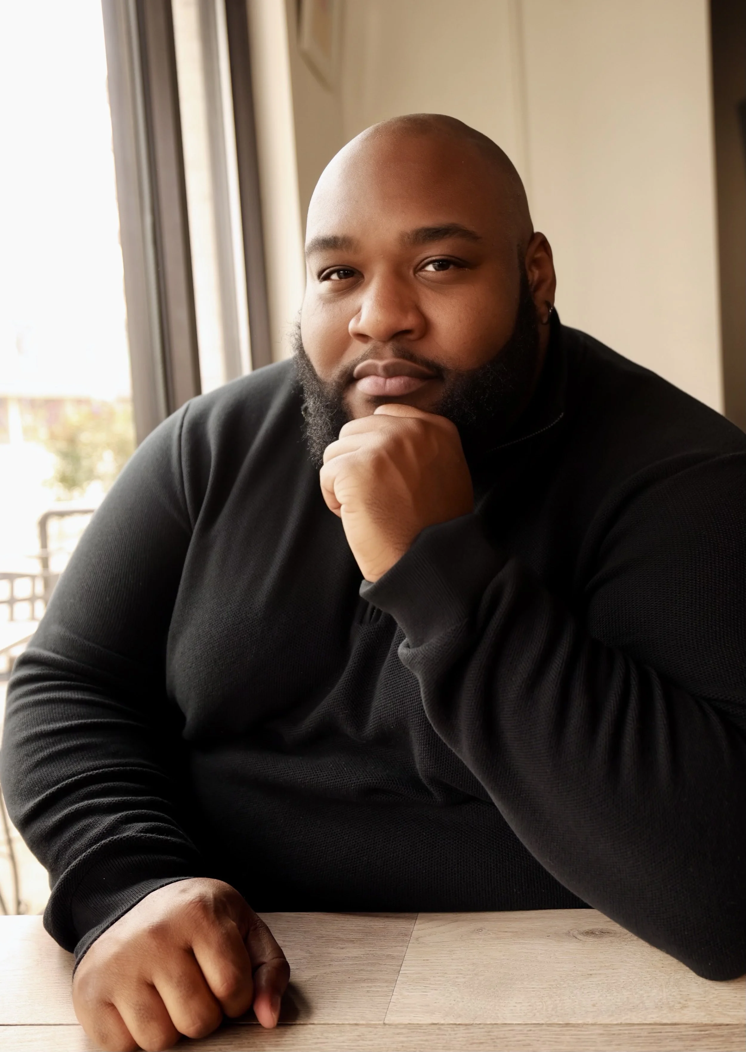 Man sitting at a table near a window in a black shirt, resting his chin on his hand.