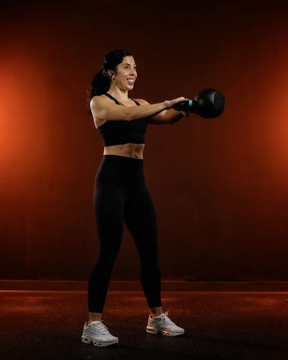 Woman in athletic wear smiling while holding a kettlebell in a gym with dark red lighting.