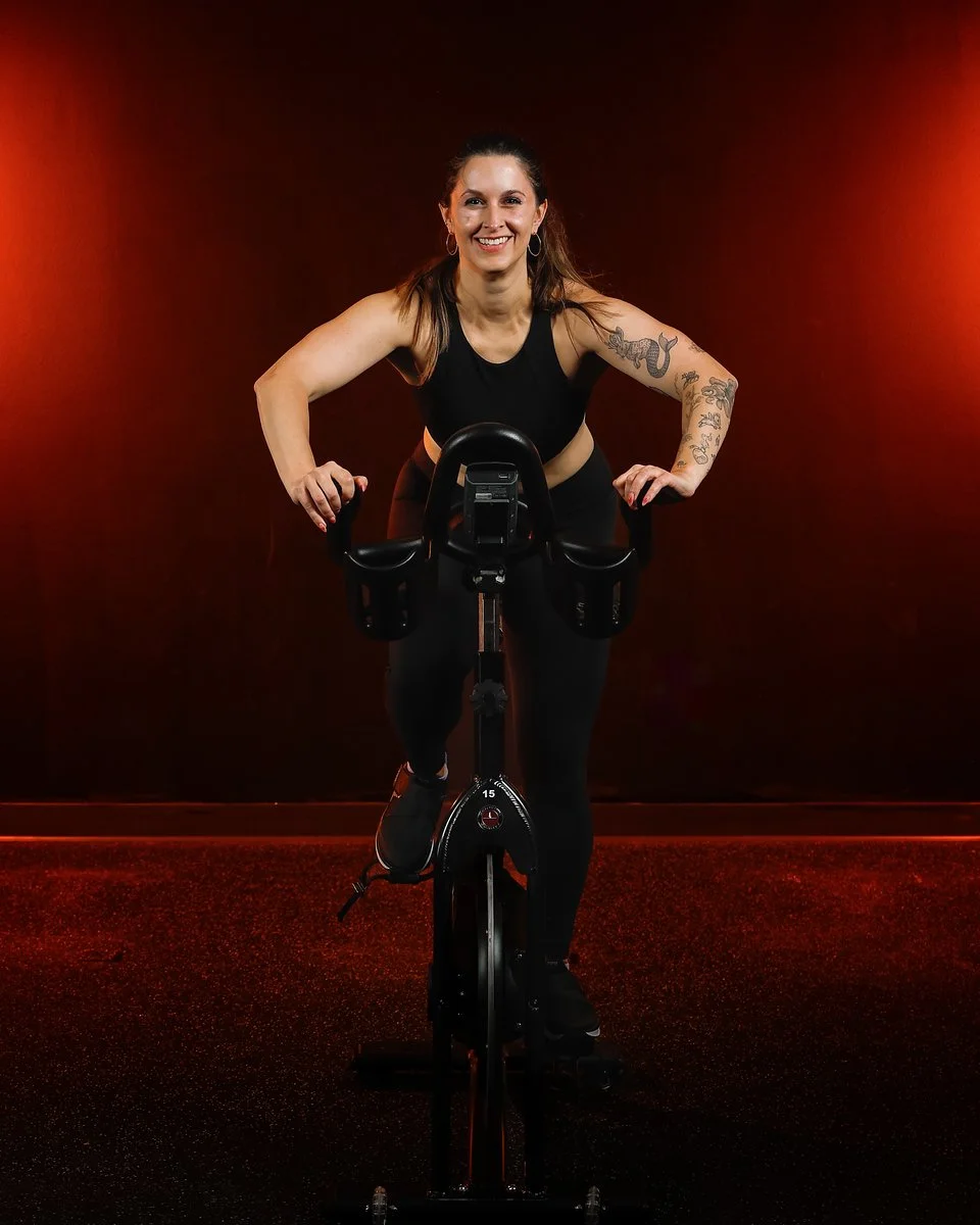 Woman smiling while riding a stationary exercise bike in a dark gym with red lighting.