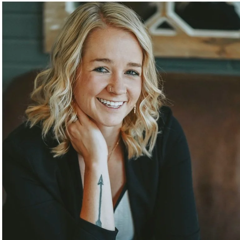 Smiling woman with blonde wavy hair, wearing a black jacket, white top, and hoop nose ring, displaying a forearm tattoo of an arrow.