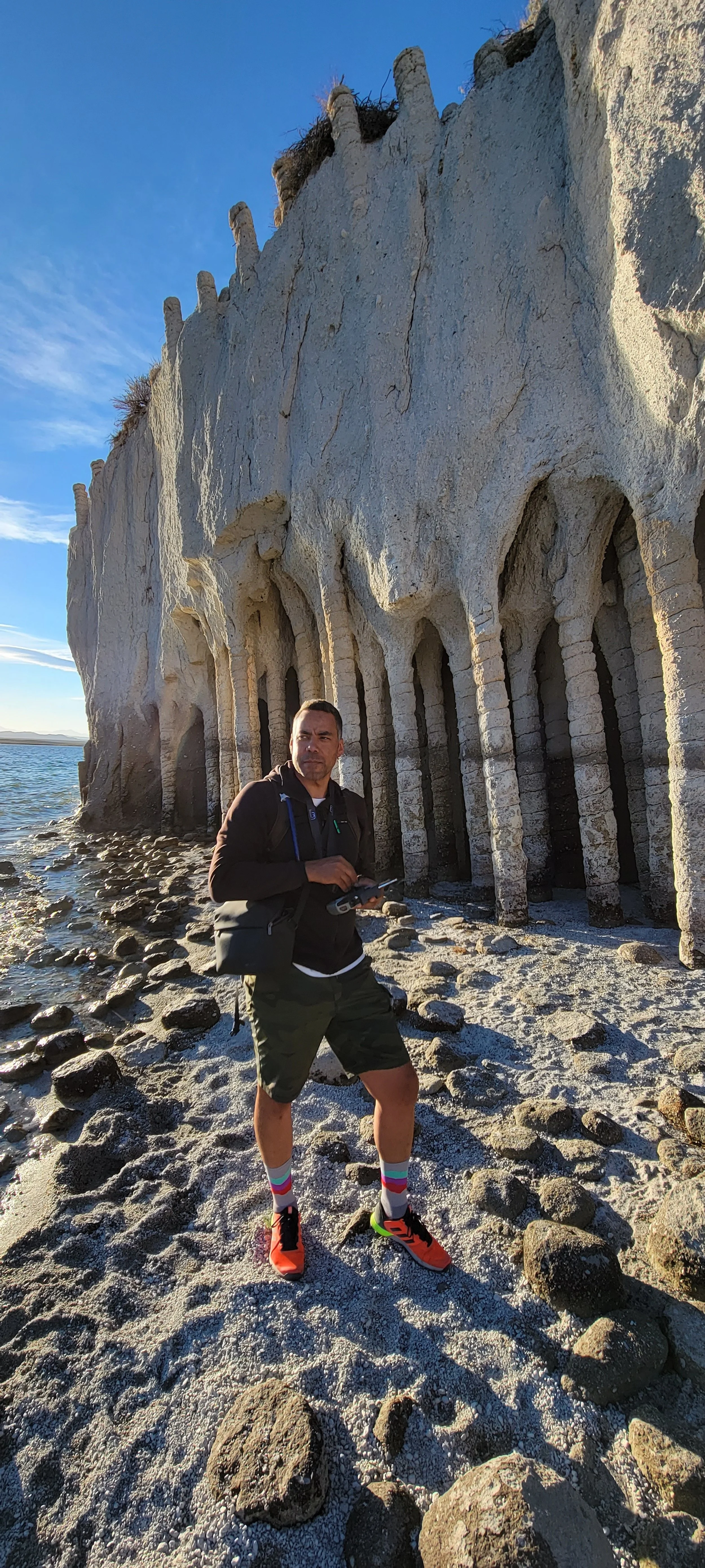 A man standing on a rocky beach next to a large cliff with a series of small arches at its base, under a clear blue sky.