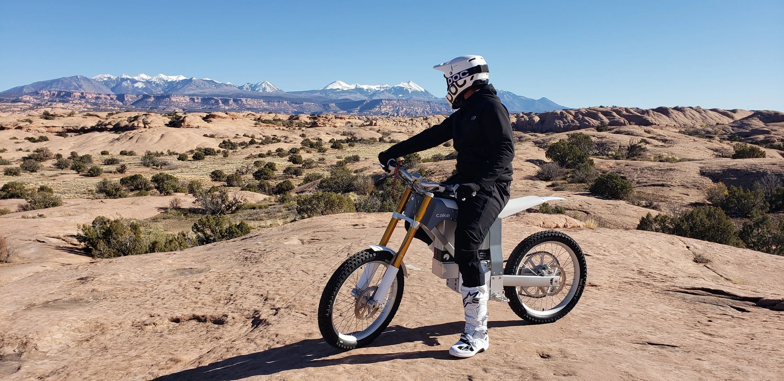 Person in black riding an electric dirt bike in a desert landscape with mountains in the background and snow-capped peaks under a clear blue sky.