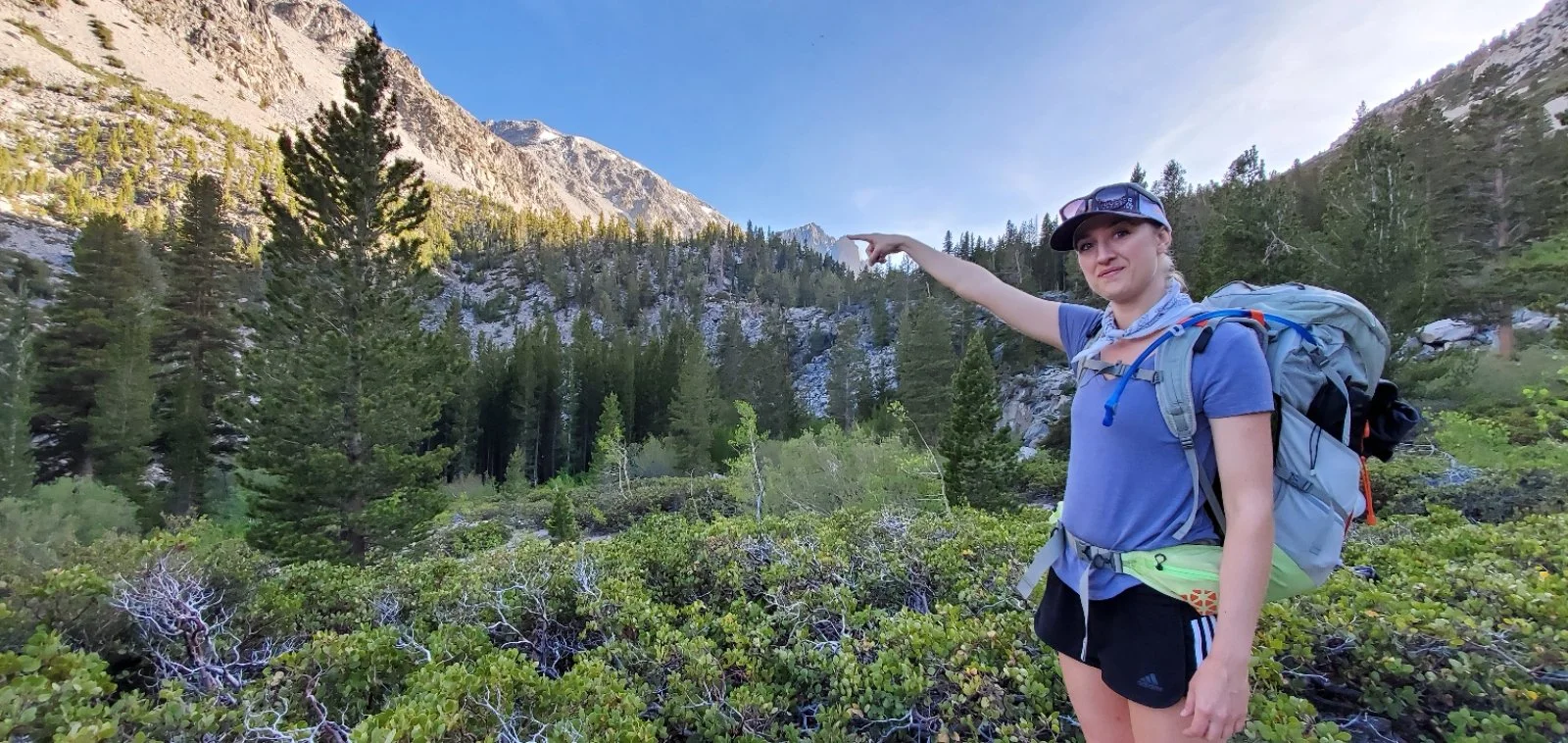 A woman hiking in a mountainous forest pointing towards the distance, wearing a blue shirt, black shorts, a cap, sunglasses, and carrying a large backpack.