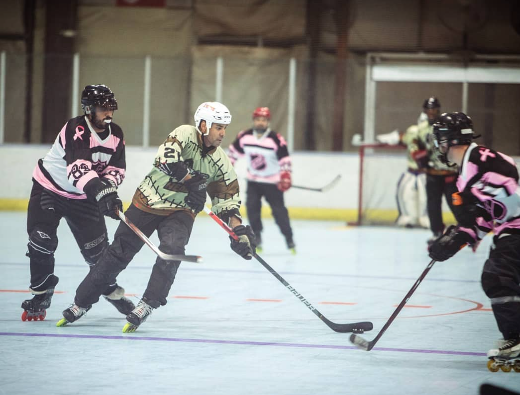 Hockey players competing on an indoor rink, with the goalie and another player in the background.