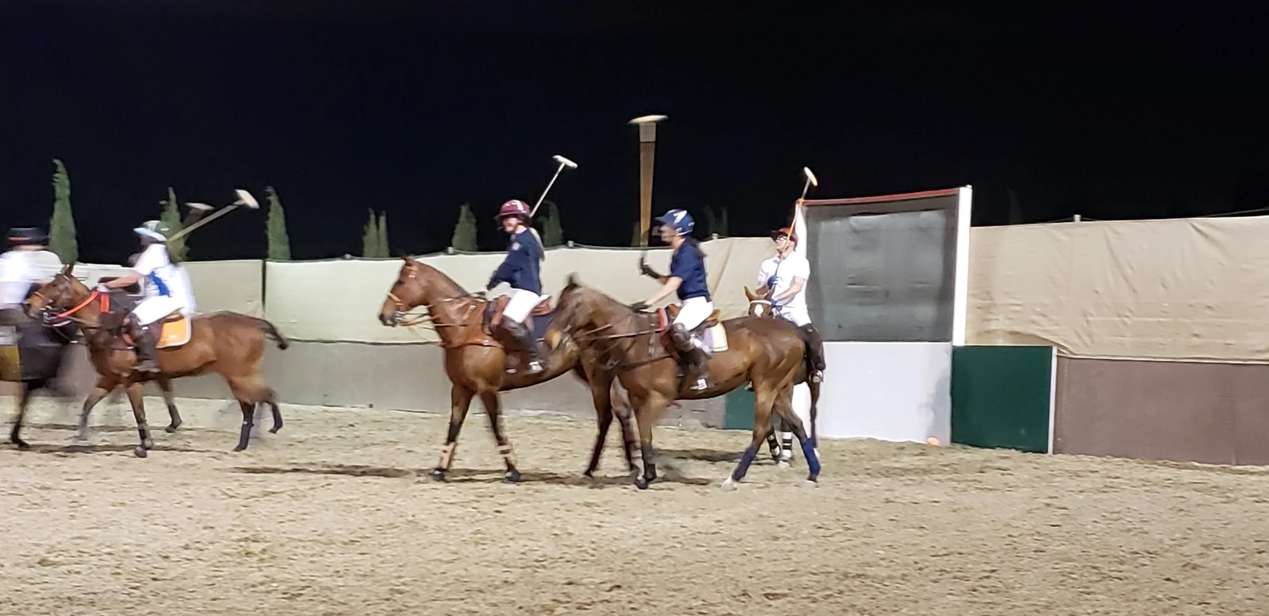 Young polo players riding horses on a field at night, wearing helmets and uniforms, with a team playing polo.