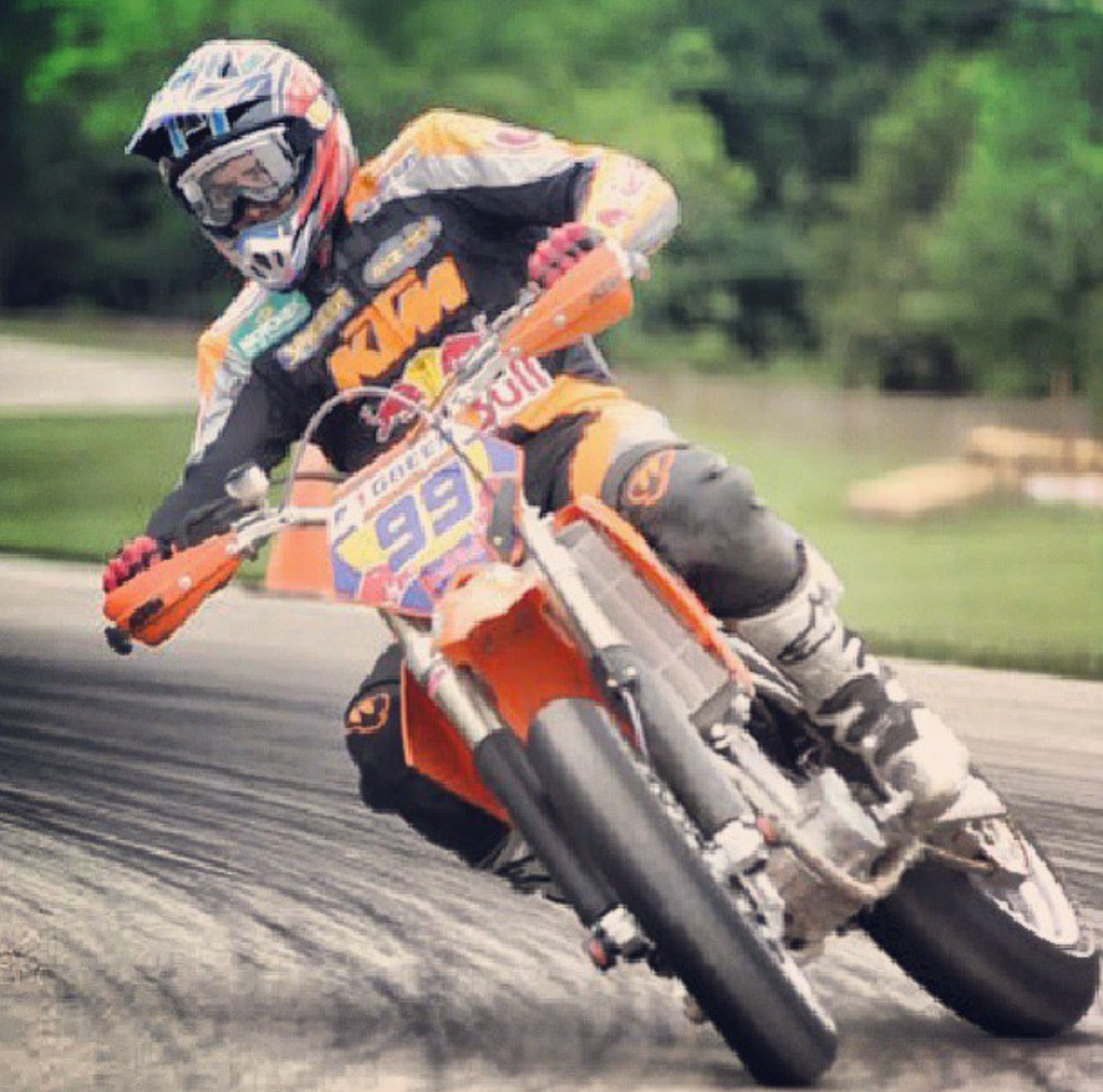 Motorcycle racer wearing a helmet and riding orange KTM dirt bike on a dirt track with greenery in the background.