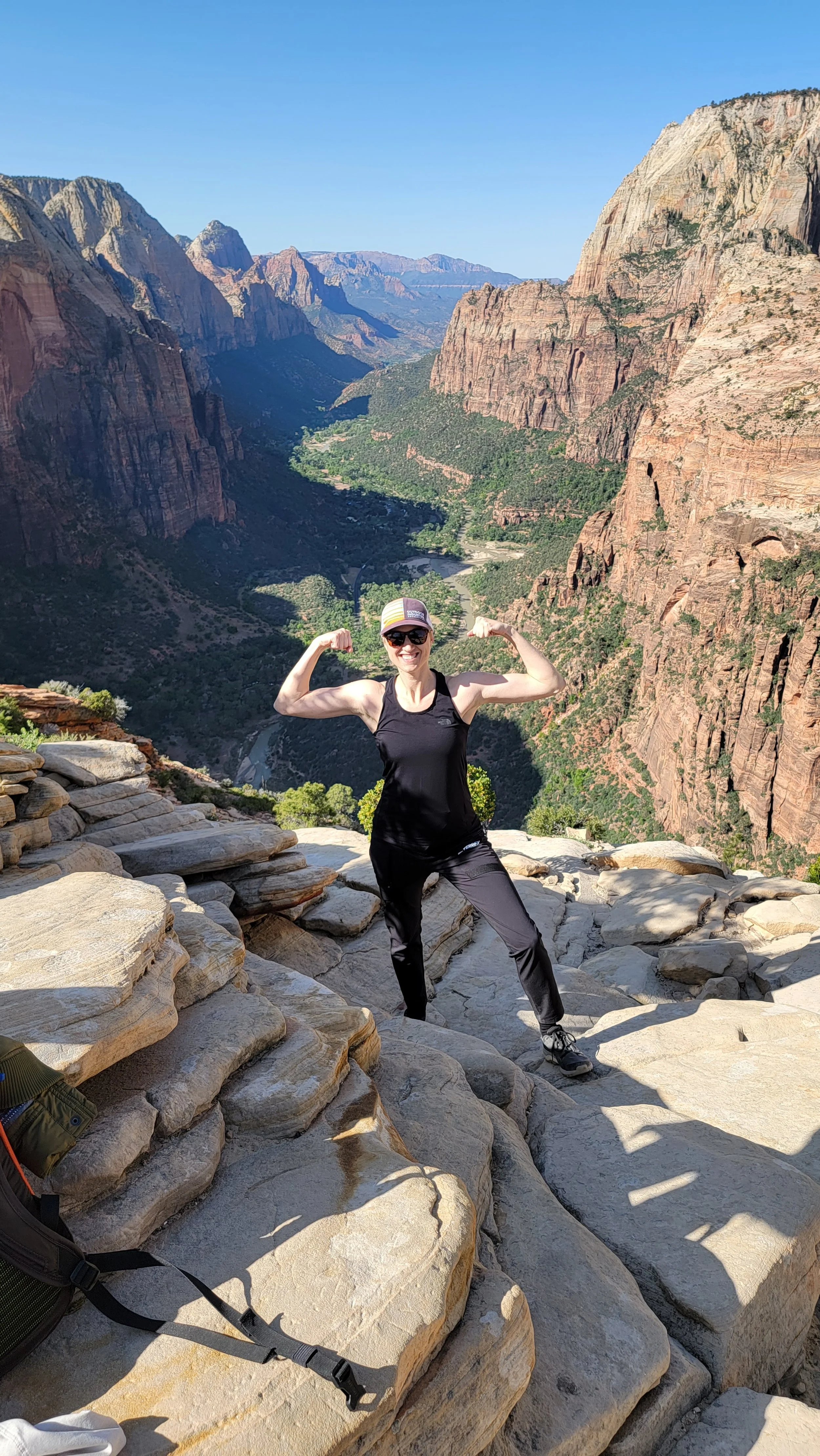 Woman in black athletic clothing and sunglasses flexing her biceps on a rocky ledge with a scenic canyon and river in the background.